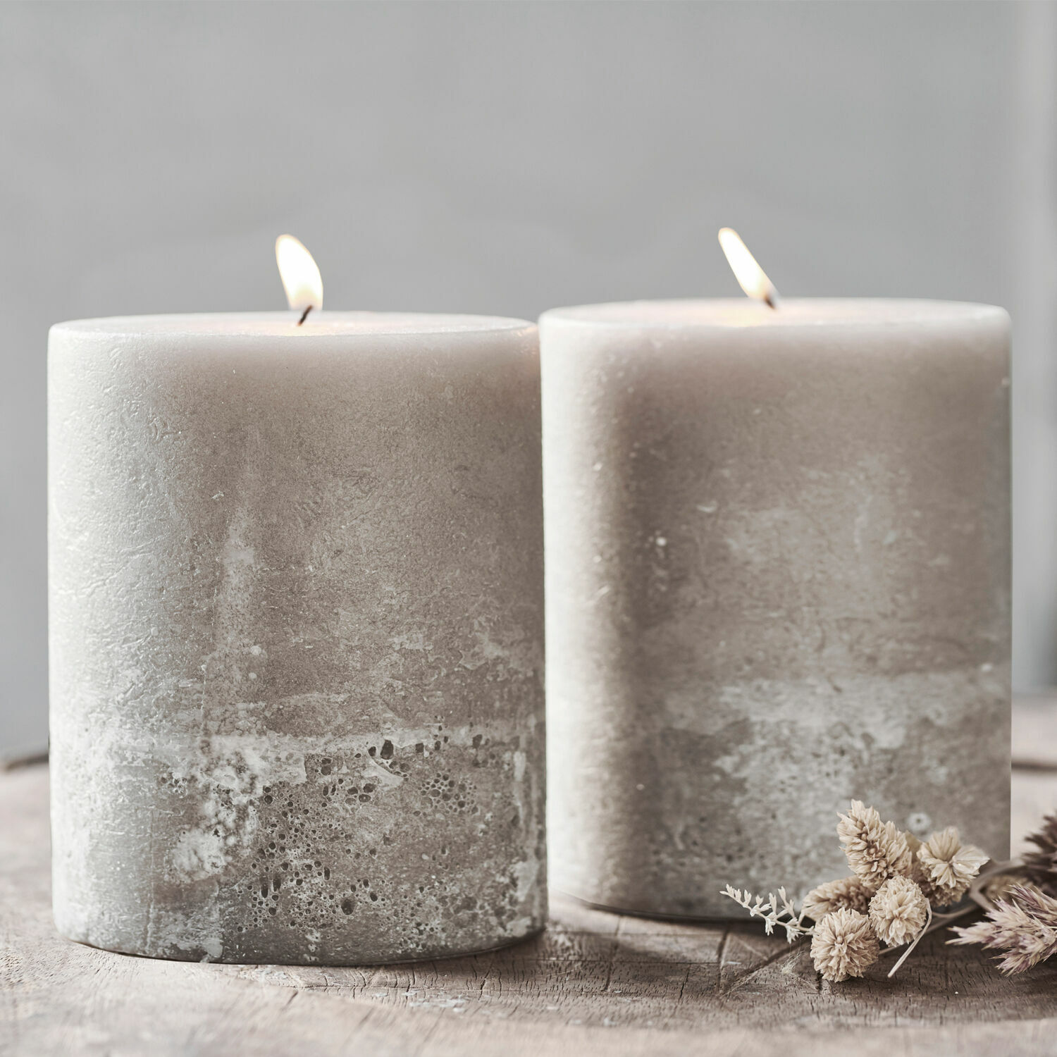 Lot de 2 bougies Oviedo Deux bougies grises en cire avec texture rustique sur une table en bois avec fleurs séchées