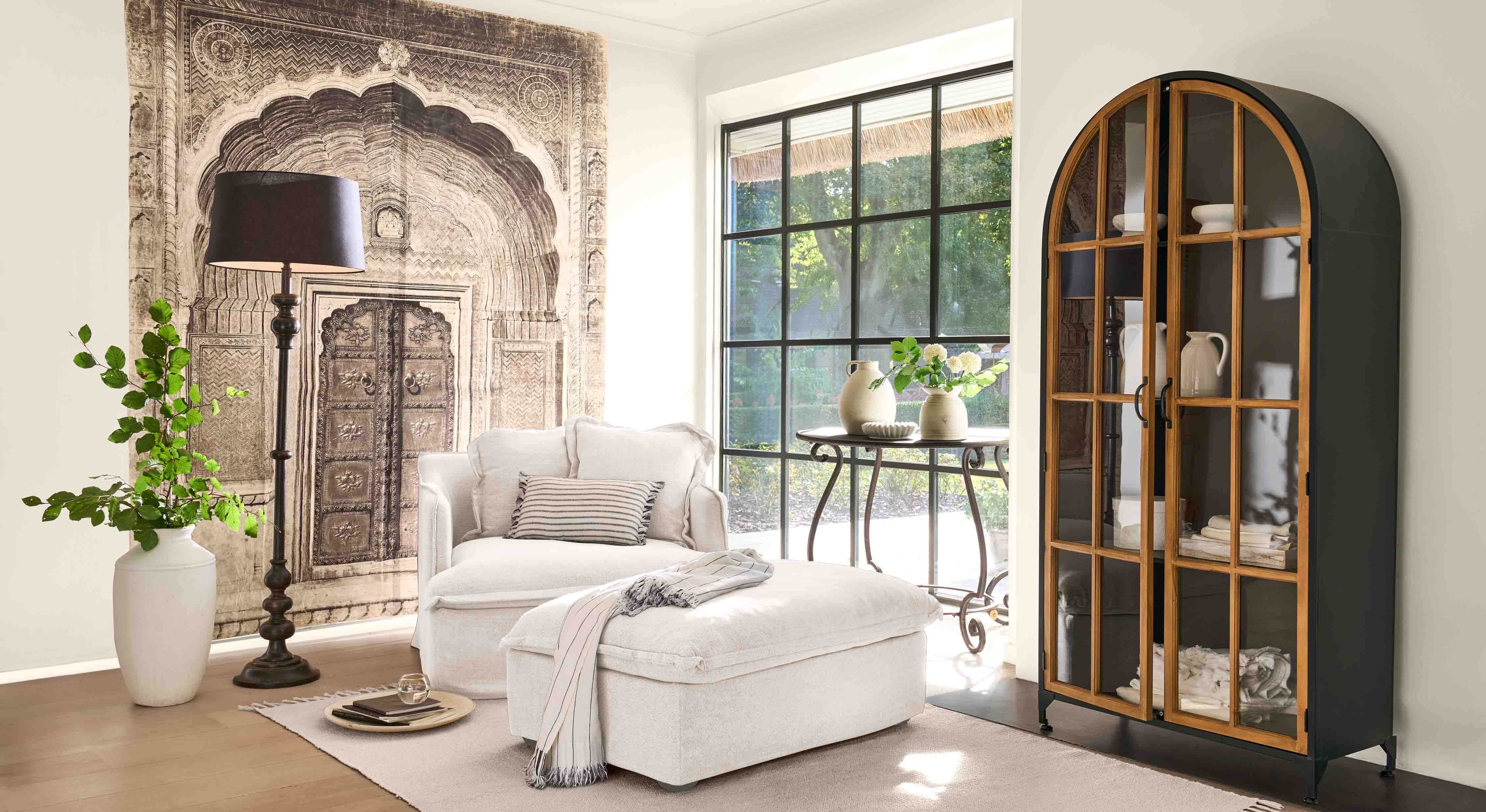 Comfortabele witte fauteuil met voetenbank en zwarte staande lamp in lichte woonkamer met decoratieve kast