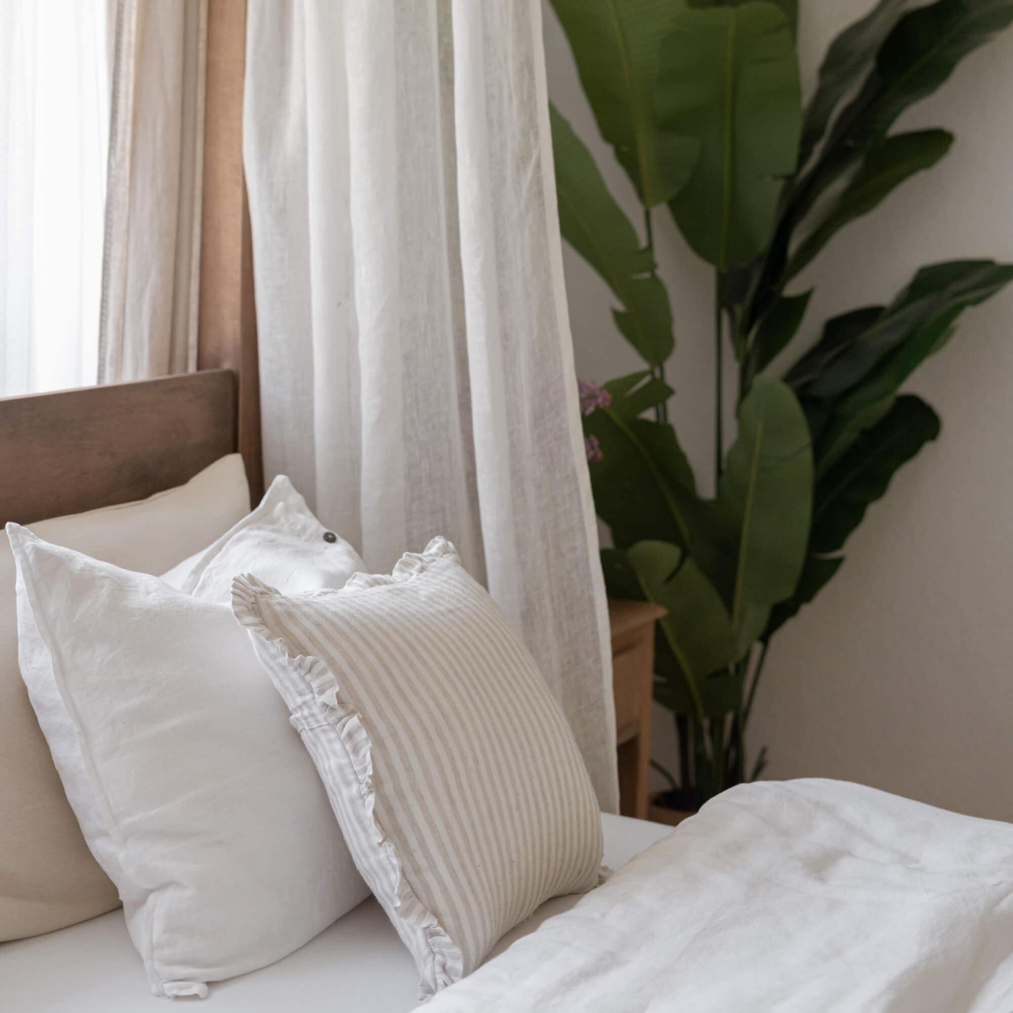 Coussins en lin blanc et beige sur un lit avec tête de lit en bois et plante verte en arrière-plan