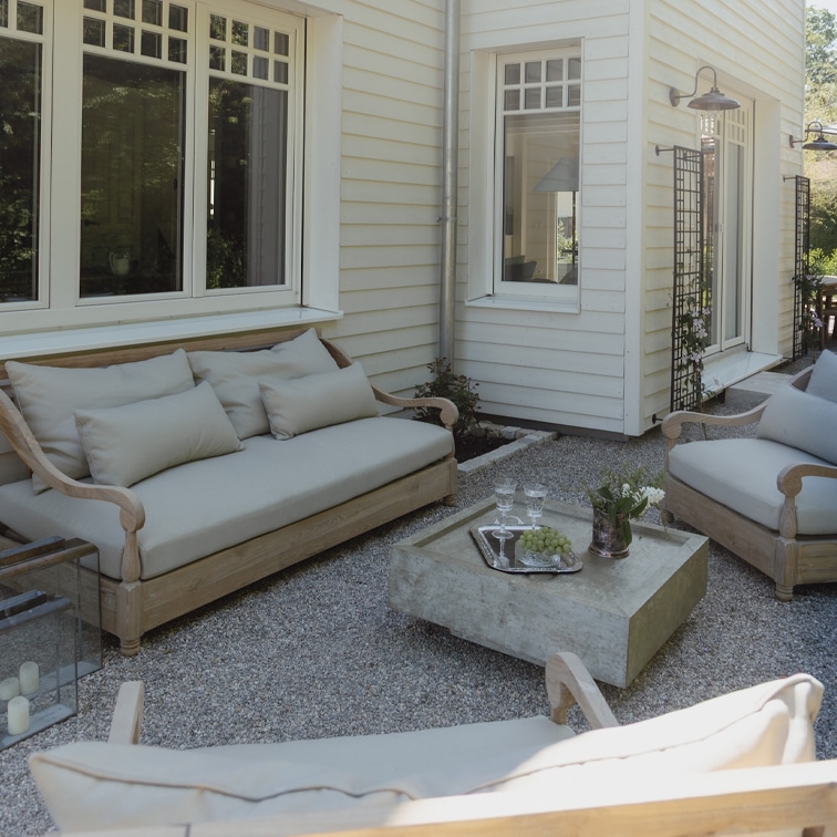 Ensemble de mobilier de jardin en bois clair avec coussins gris sur patio en gravier devant une maison blanche