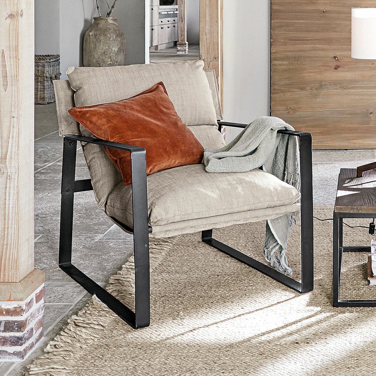 Fauteuil Seuvard Beige stoffen fauteuil met zwart metalen frame, oranje kussen en grijze deken in woonkamer