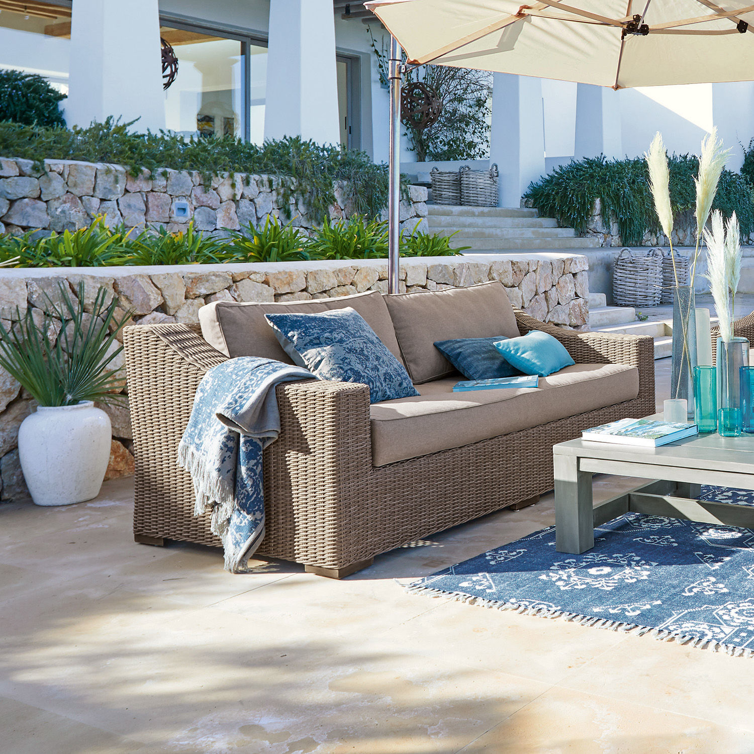 Canapé d'extérieur en rotin beige avec coussins bleus et parasol sur terrasse ensoleillée