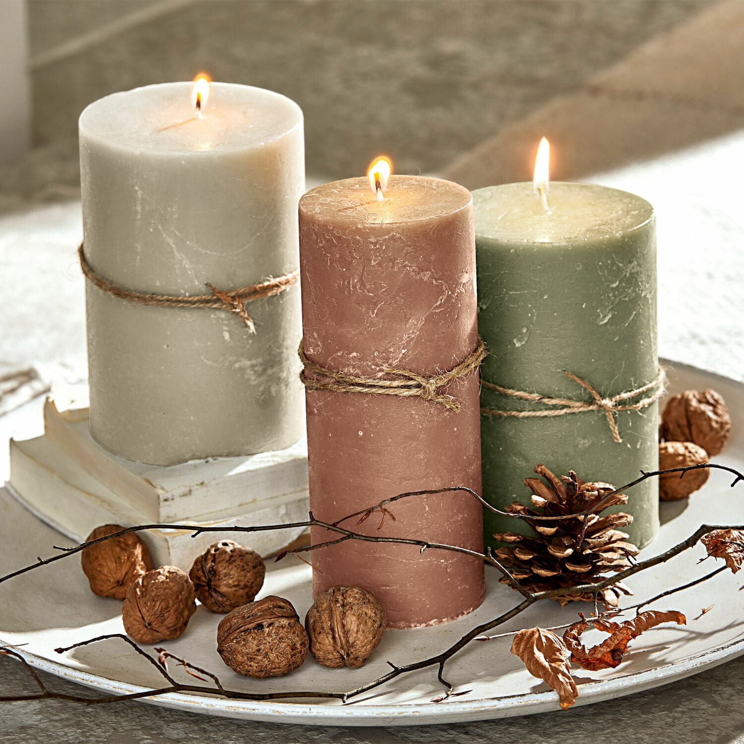 Lot de 3 bougies Salis Trois bougies en cire allumées beige, rose et vert sur un plateau décoratif avec des noix et des branches