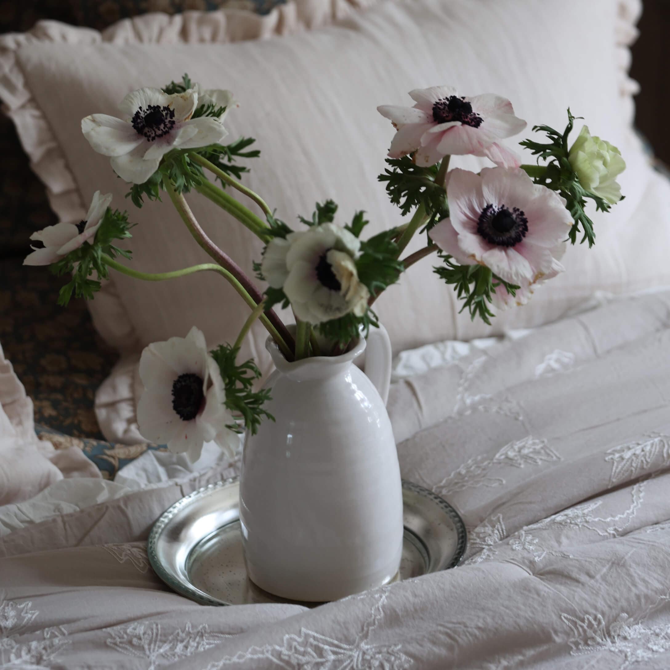 Witte keramische kan met delicate witte anemonen op beige geborduurd beddengoed