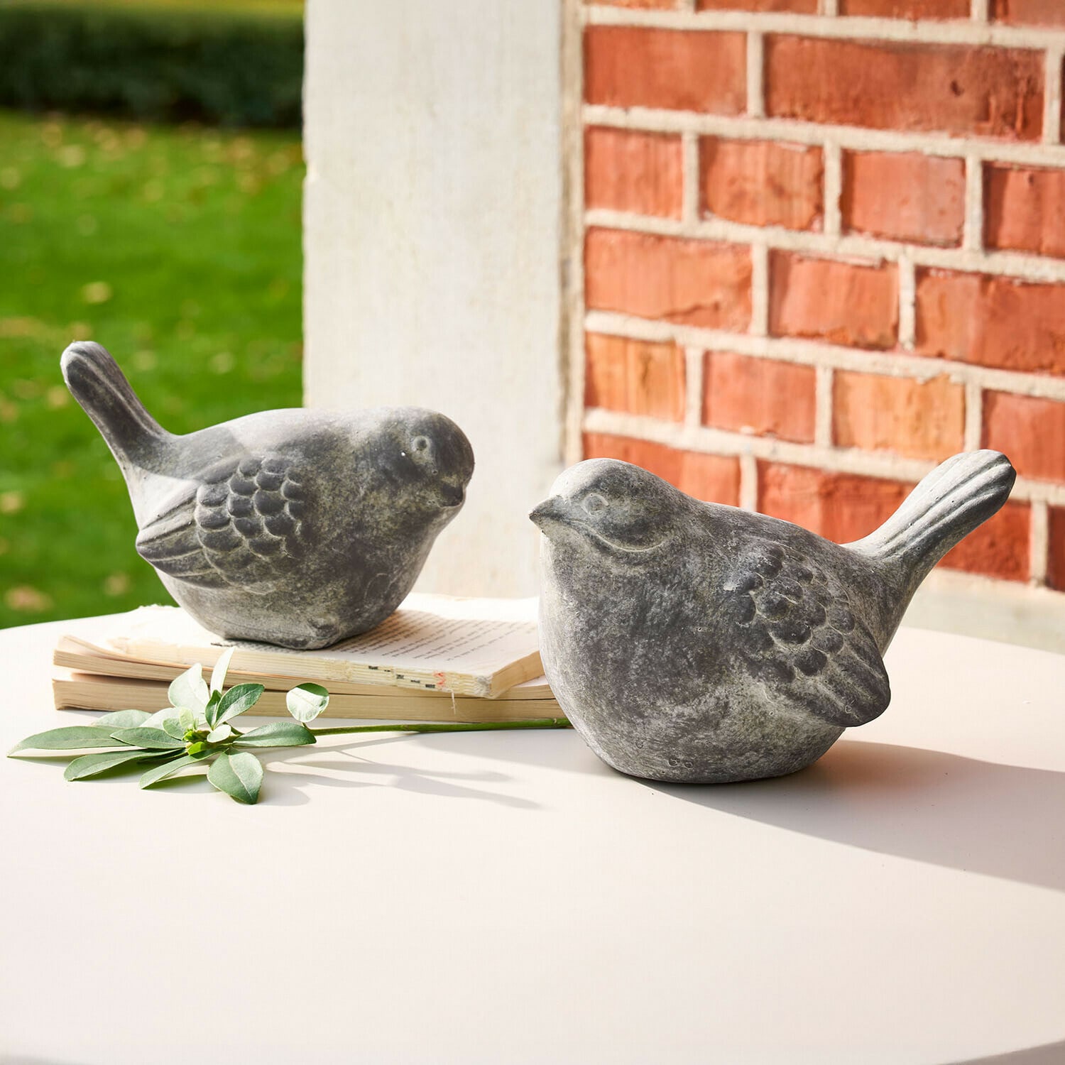 Lot de 2 oiseaux décoratifs Haldoríe Deux figurines d'oiseaux décoratives en pierre grise sur une table blanche avec un livre et une feuille verte