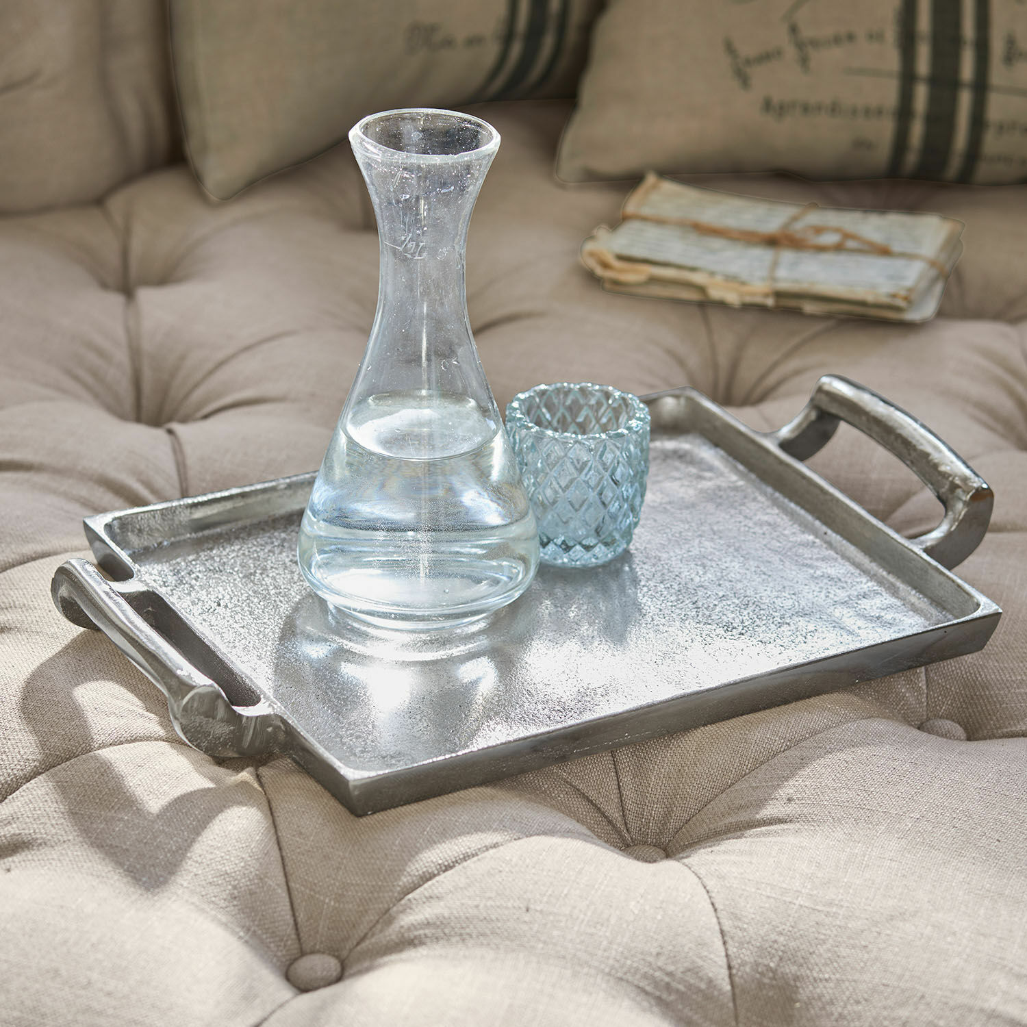 Plateau Connaire Plateau décoratif en métal argenté posé sur un tissu beige matelassé avec carafe et verre en verre