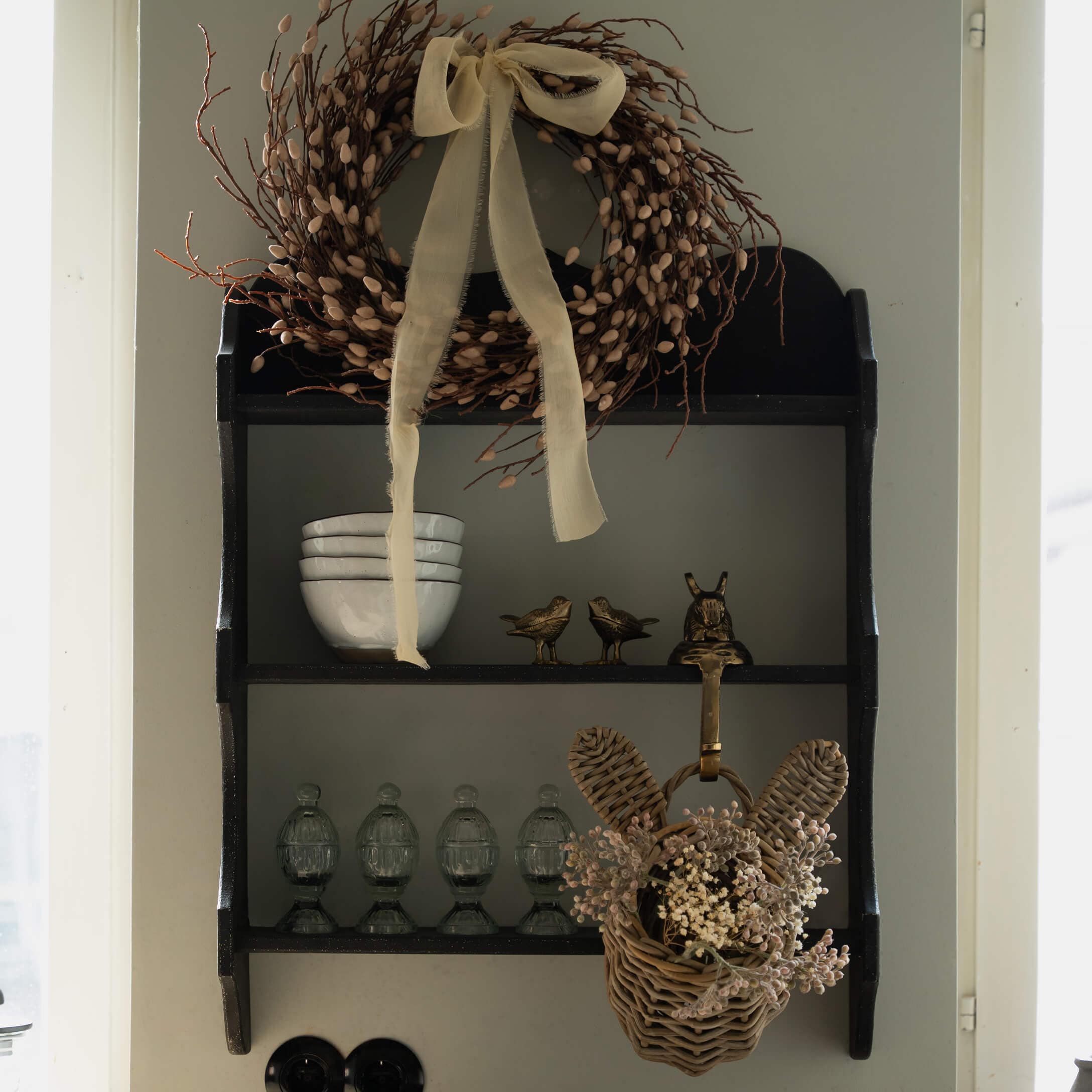 Zwart houten wandrek met decoratieve krans en rieten mand met gedroogde bloemen eraan