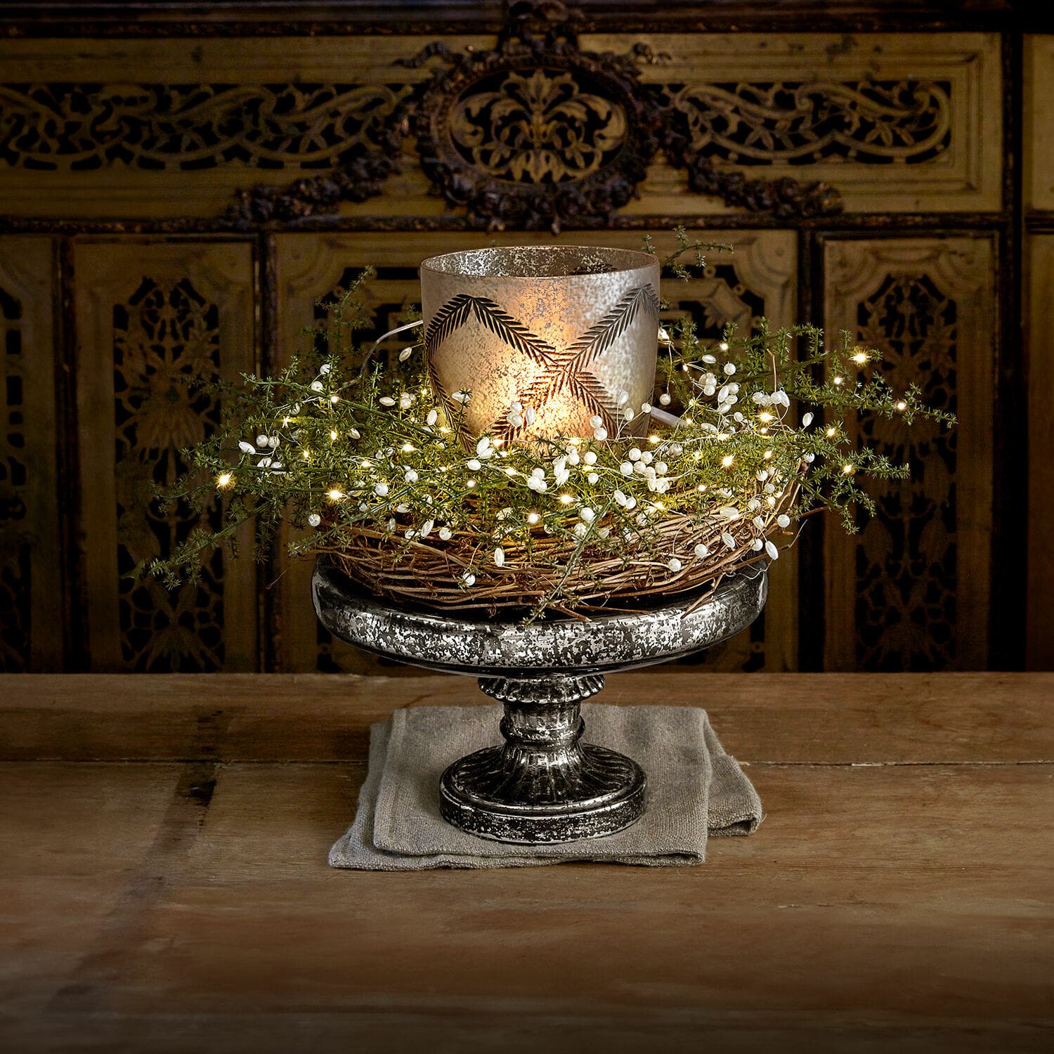 Présentoir à étage Incantesse Bougeoir décoratif en métal argenté avec couronne en osier et guirlande lumineuse LED sur table en bois