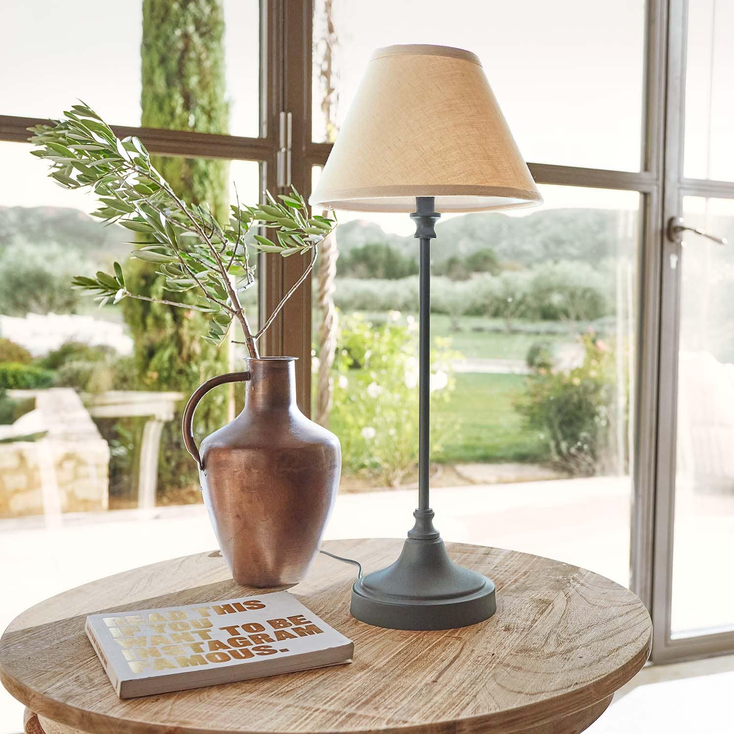 Pichet en métal couleur cuivre avec branche et lampe de table à abat-jour beige sur table en bois