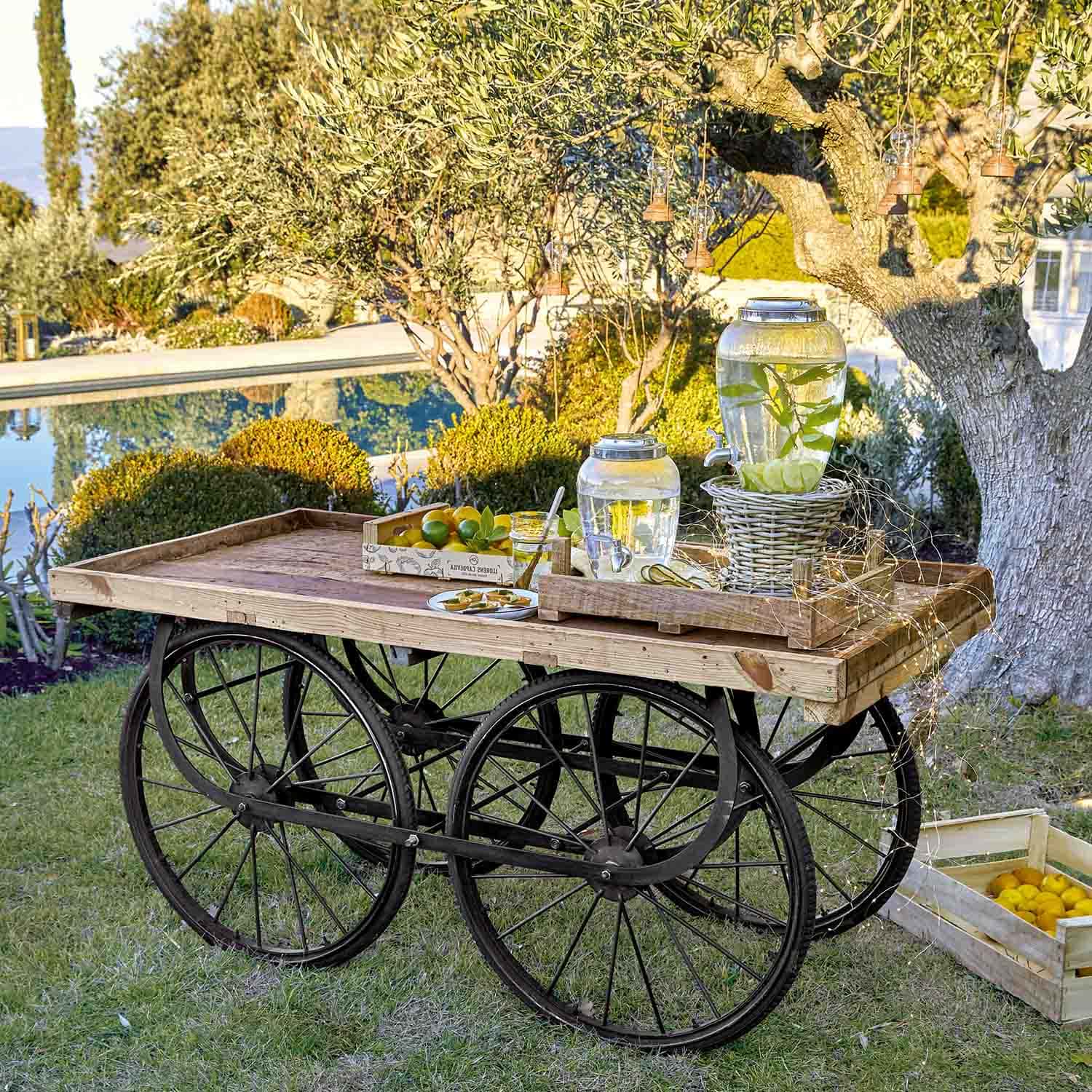 Chariot en bois avec roues noires portant des distributeurs d'eau en verre citronnée dans un jardin