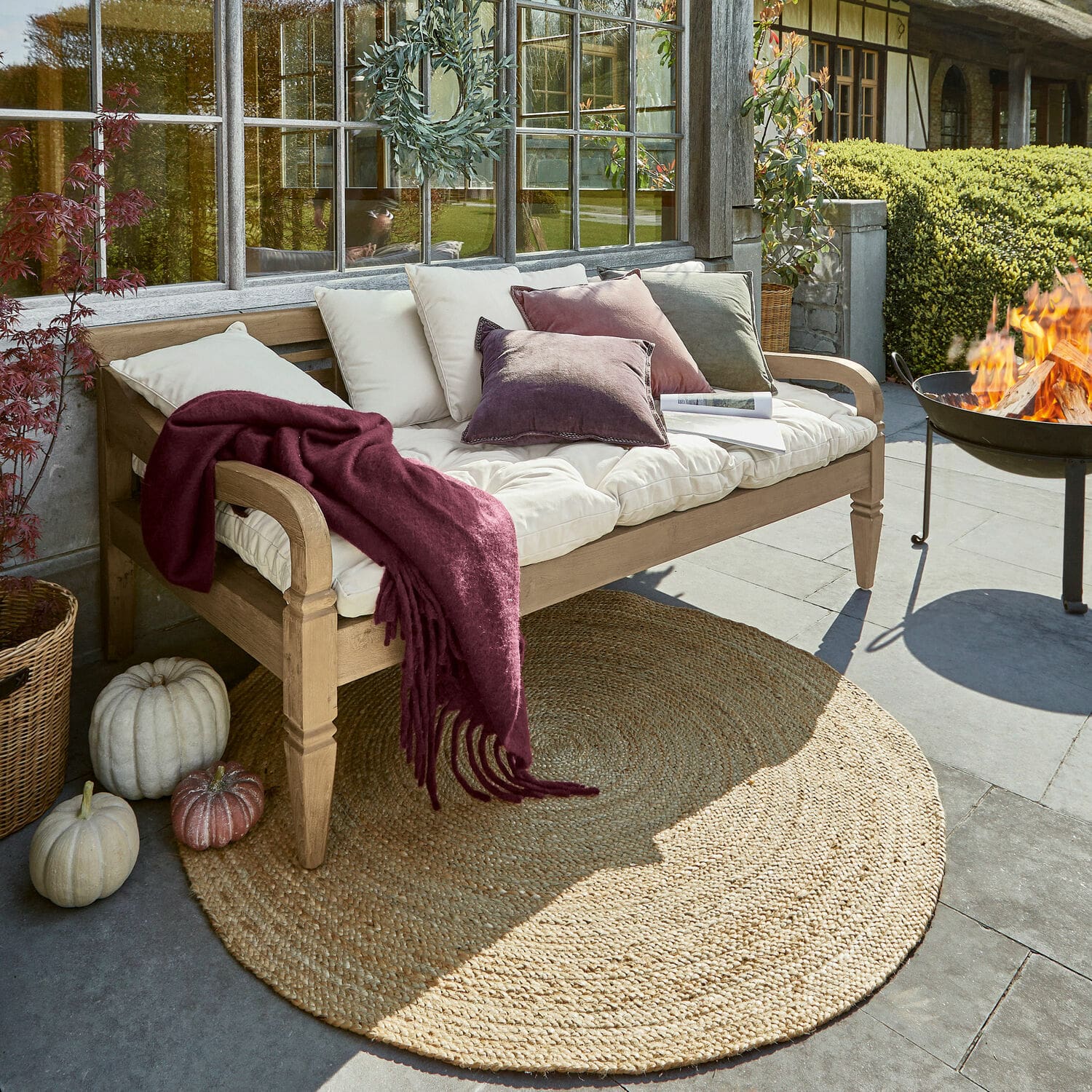 Banc de jardin en bois avec coussins crème et plaid bordeaux sur terrasse avec tapis en jute