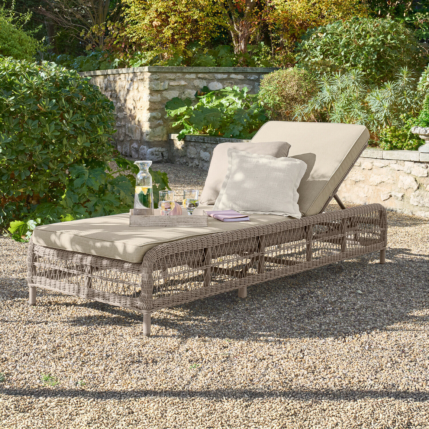 Chaise longue de jardin beige avec coussin et structure en osier sur sol en gravier dans un jardin verdoyant