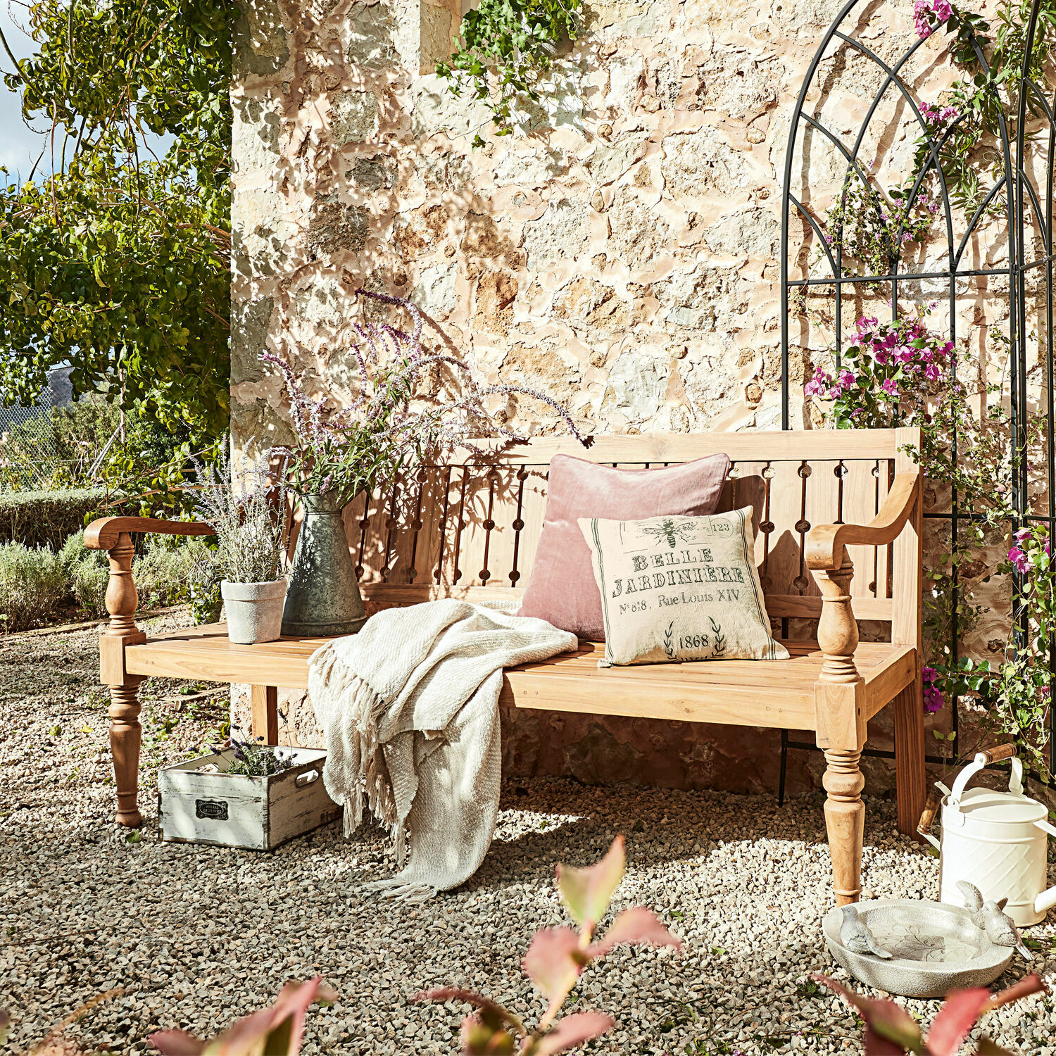Banc Loudoun Banc de jardin en bois clair avec coussins et plaid sur patio en gravier entouré de plantes