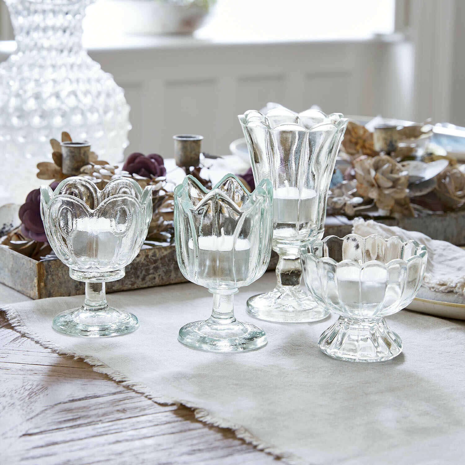 Lot de 4 photophores Amisette Ensemble de quatre bougeoirs en verre transparent de formes variées sur une nappe claire
