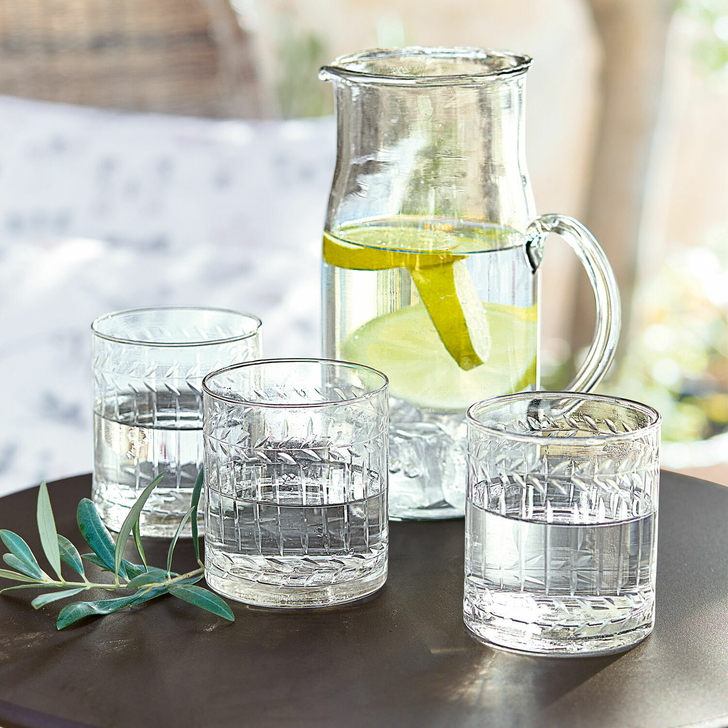 Lot de 6 verres Padiès Carafe en verre avec tranches de citron et trois verres d'eau sur une table sombre avec feuillage décoratif