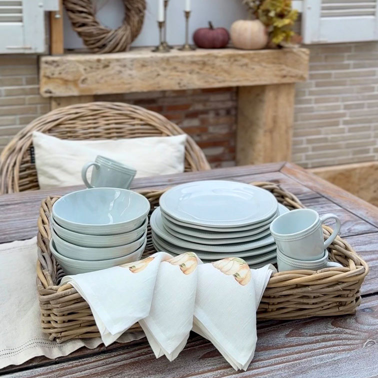 Set lichtgrijs keramisch servies met borden, kommen en mokken in een rieten mand op een houten tafel