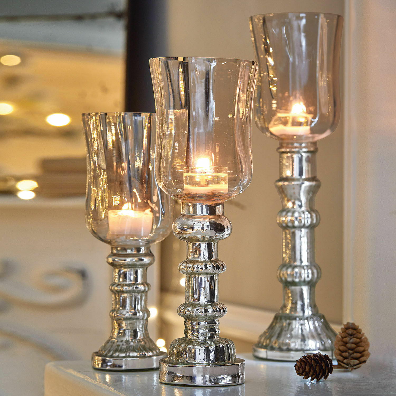 Lot de 3 photophores Fréjeville Ensemble de trois bougeoirs argentés en verre et métal avec bougies allumées et pommes de pin décoratives