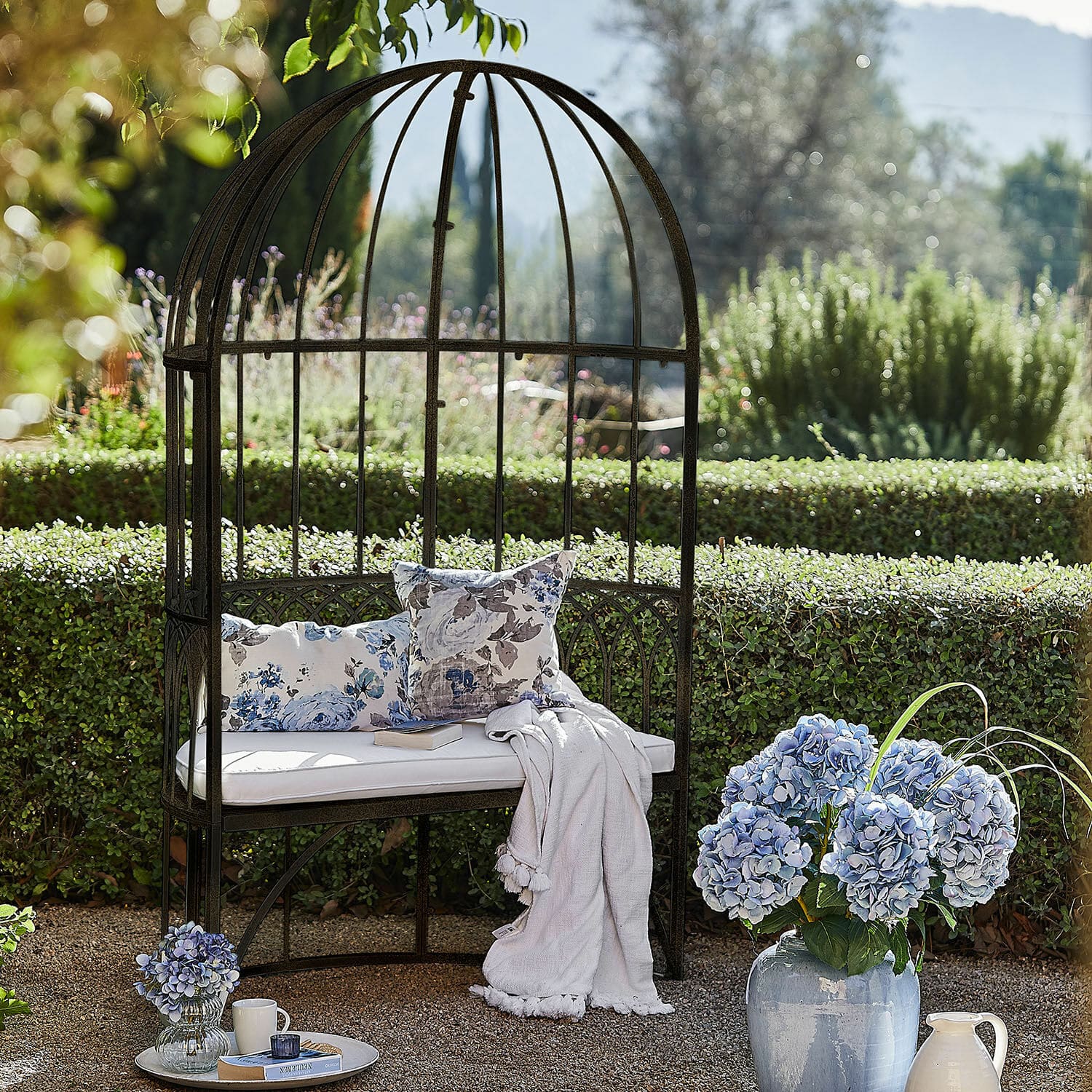 Banc Rivandelle Jardin avec banc en métal en forme de cage, coussin blanc et fleurs bleues dans un vase