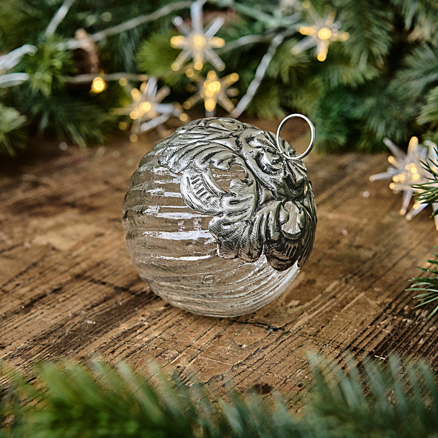 Boule de Noël Zoriline Boule de Noël en verre avec ornement métallique argenté posée sur une table en bois entourée de branches vertes
