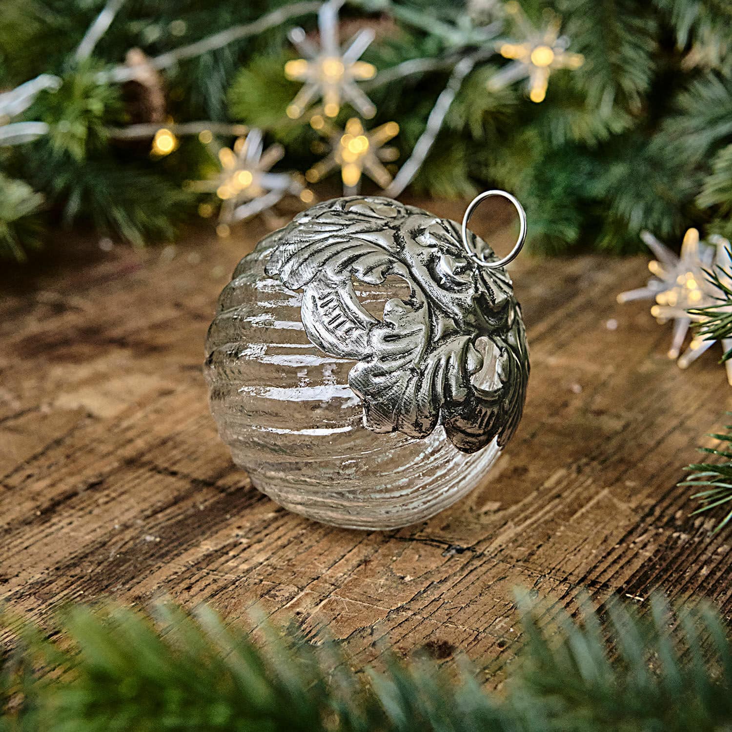 Boule de Noël Zoriline Boule de Noël en verre avec ornement métallique argenté posée sur une table en bois entourée de branches vertes