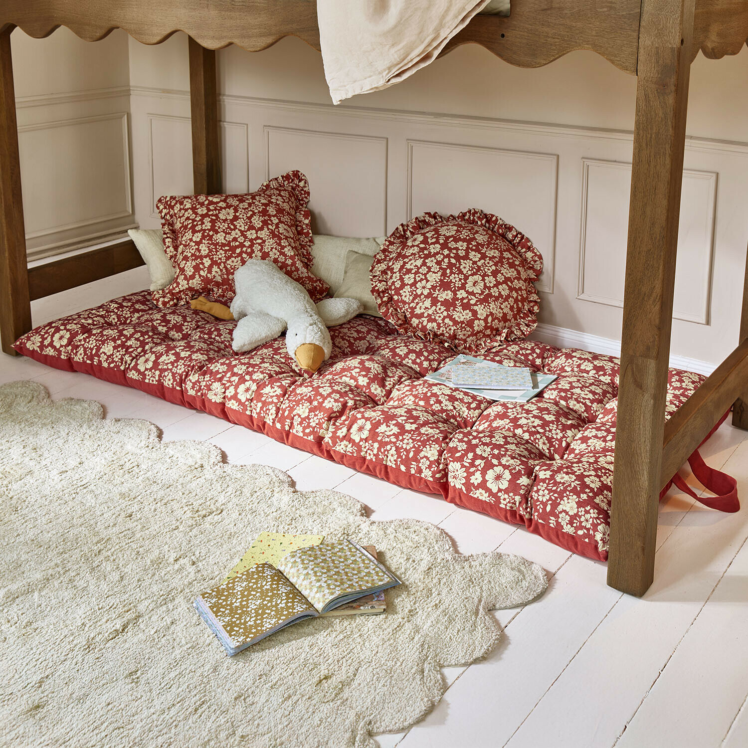 Matelas de jeu Frambelle Matelas rouge à motif floral avec coussins sous une table en bois, peluche et livres posés au sol