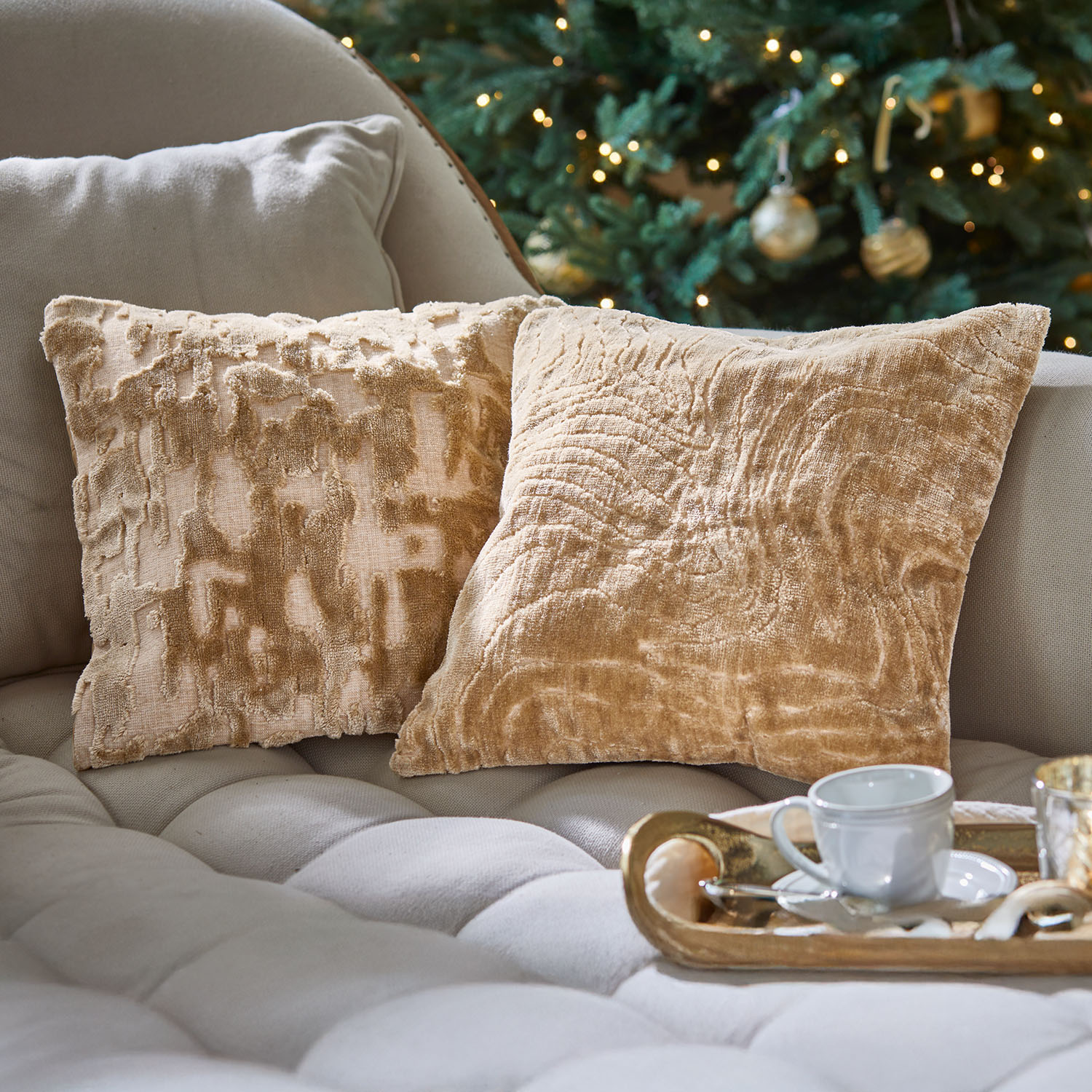 Deux coussins décoratifs beige en tissu doux sur un canapé clair près d'un sapin de Noël décoré