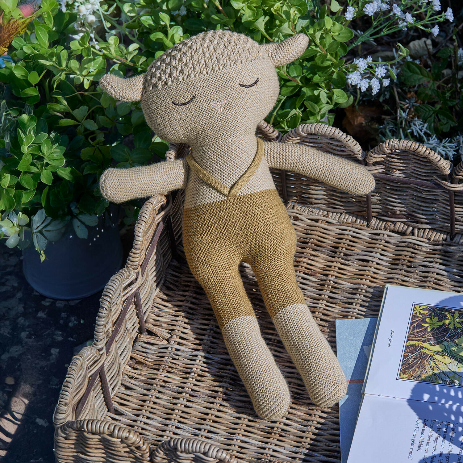 Knuffeltje Cuddlecloud Beige gebreide schapenknuffel ligt in een rieten mand naast een boek in de tuin