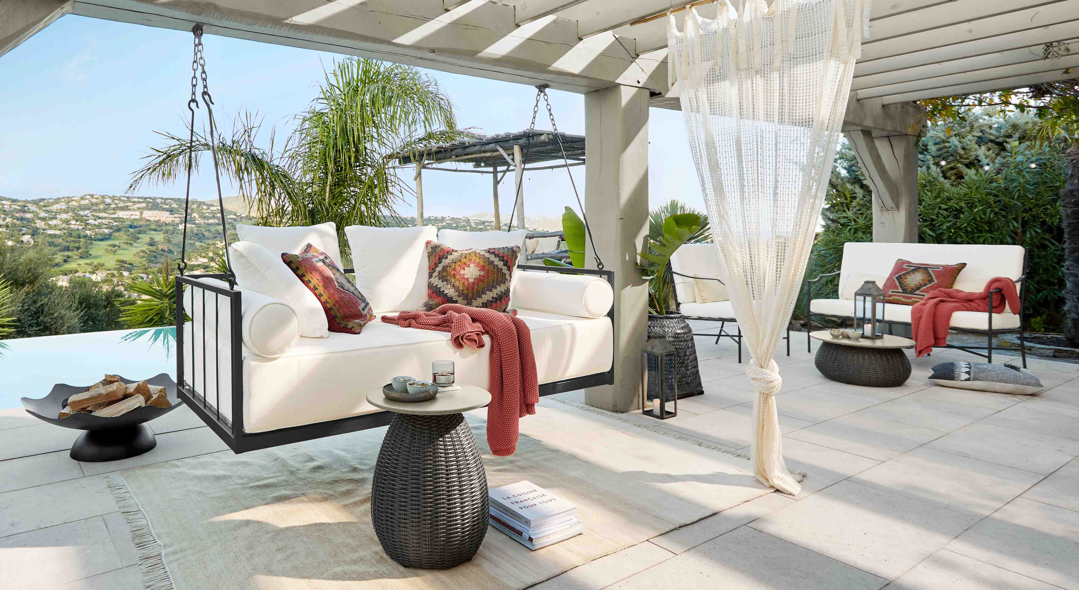 Witte hangbank met metalen frame en kleurrijke kussens op terras met uitzicht op heuvels