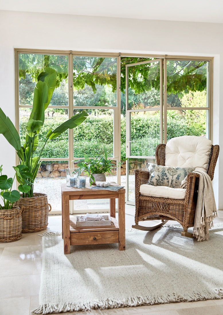 Fauteuil à bascule en osier avec coussin crème et table en bois sur tapis clair dans un intérieur lumineux