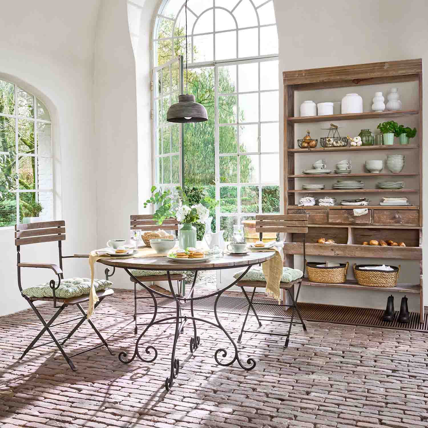 Eetkamer met ronde tafel met metalen poten en houten stoelen op bakstenen vloer bij groot raam
