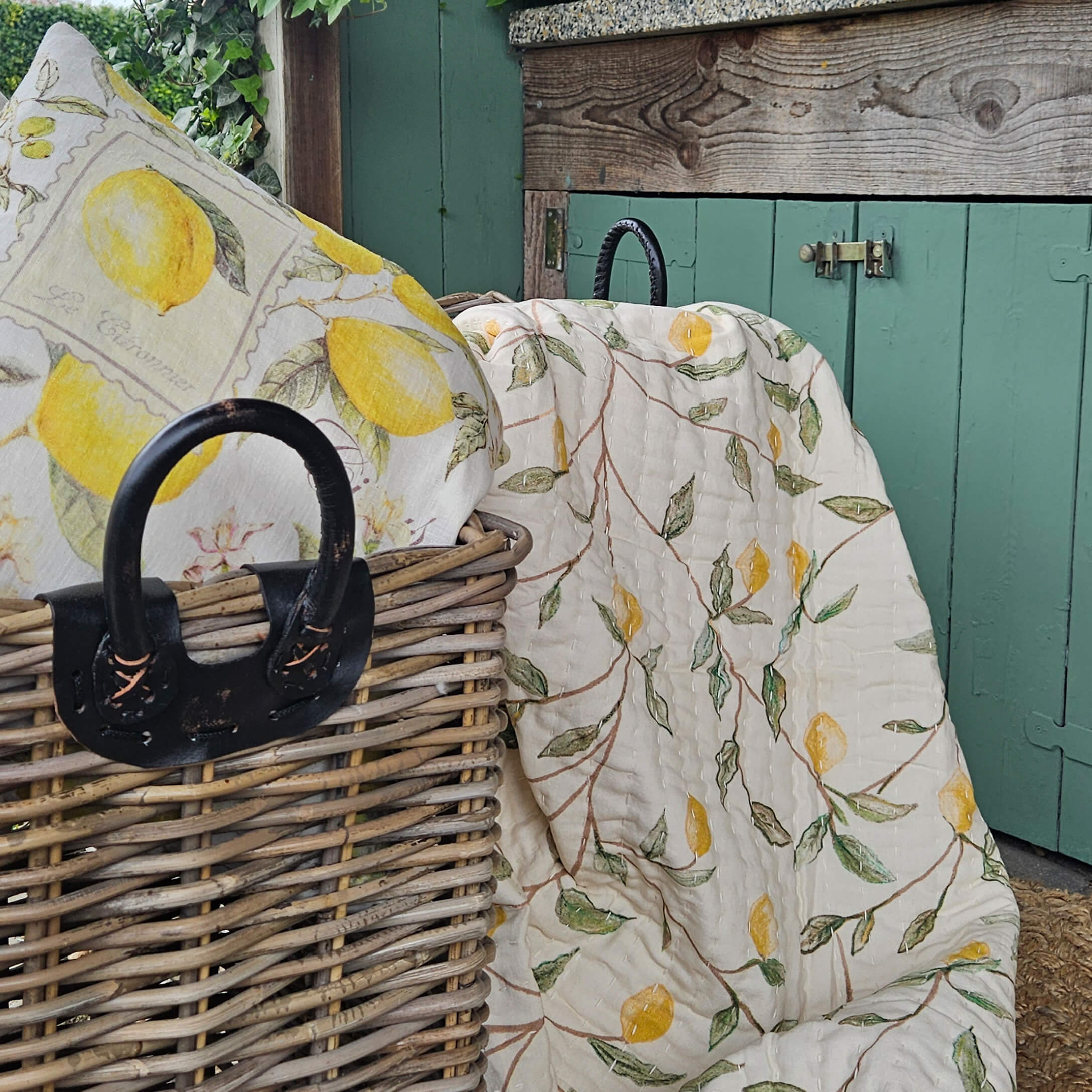 Plaid et coussin en lin avec motif citron jaune posés sur un panier en osier devant une porte verte
