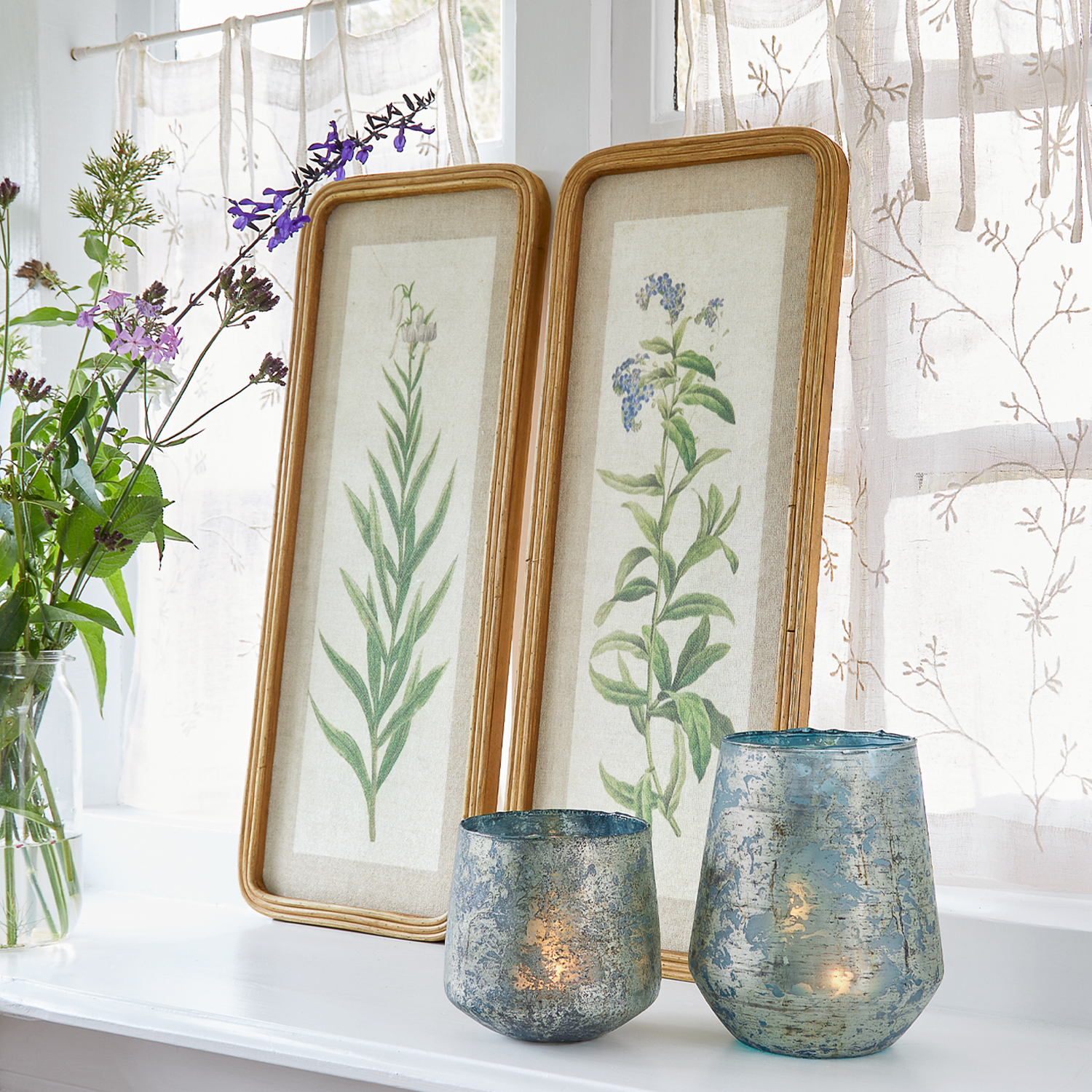 Deux cadres avec illustrations botaniques et bougeoirs en verre bleu sur un rebord de fenêtre avec rideaux