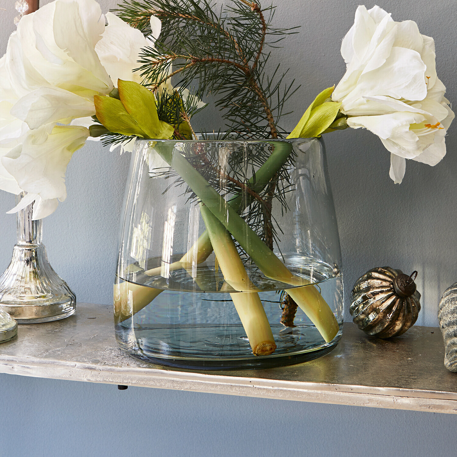 Transparante glazen vaas met witte bloemen en groene takken op een decoratieve plank