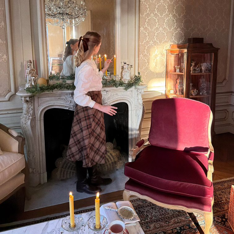 Vrouw die kaarsen aansteekt bij een marmeren open haard in een elegante woonkamer met bordeauxrode stoel