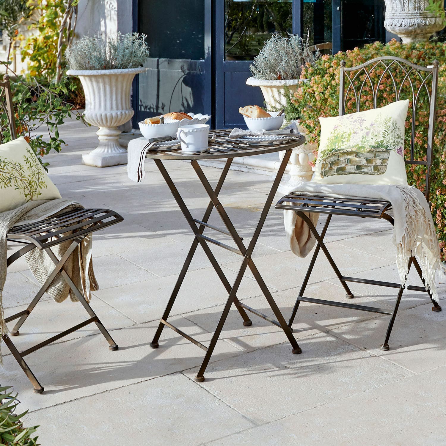 Table Brignoles Table et chaises pliantes en métal brun sur terrasse avec coussins décoratifs et vaisselle blanche