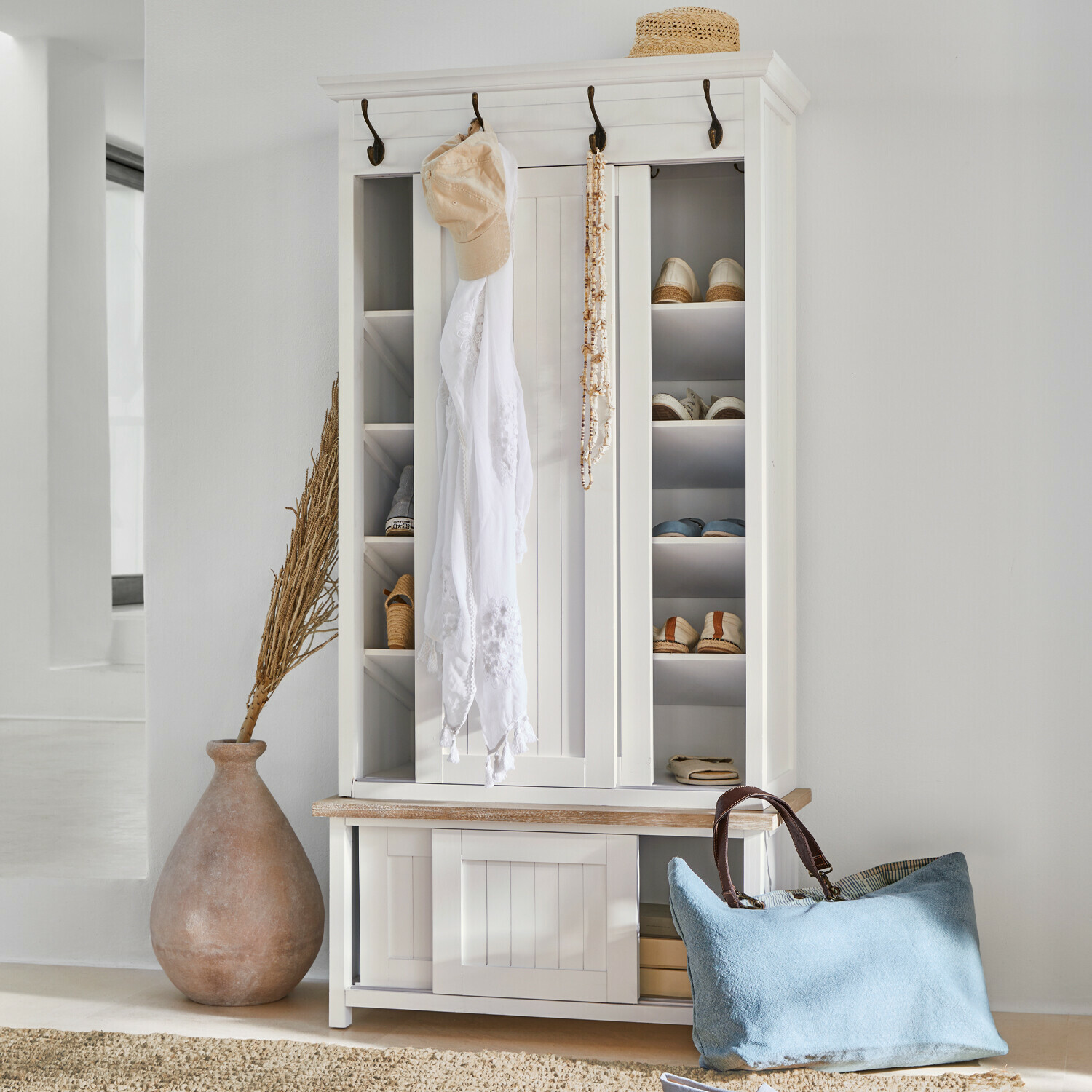 Witte houten schoenenkast met haken en planken, blauwe tas en strohoed als decoratie