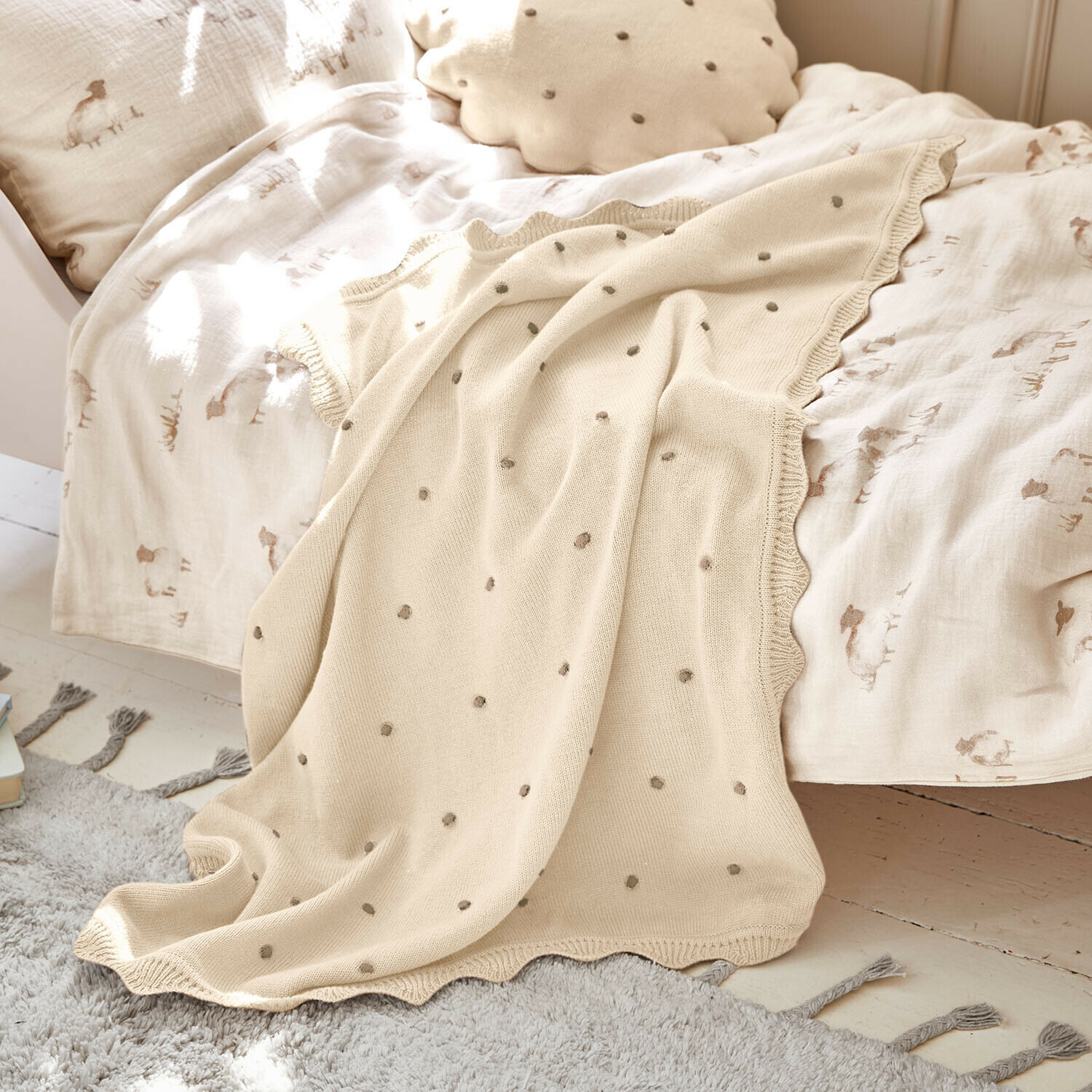 Plaid Saphie Plaid tricoté beige à pois gris sur un lit avec linge de lit clair et coussin naturel