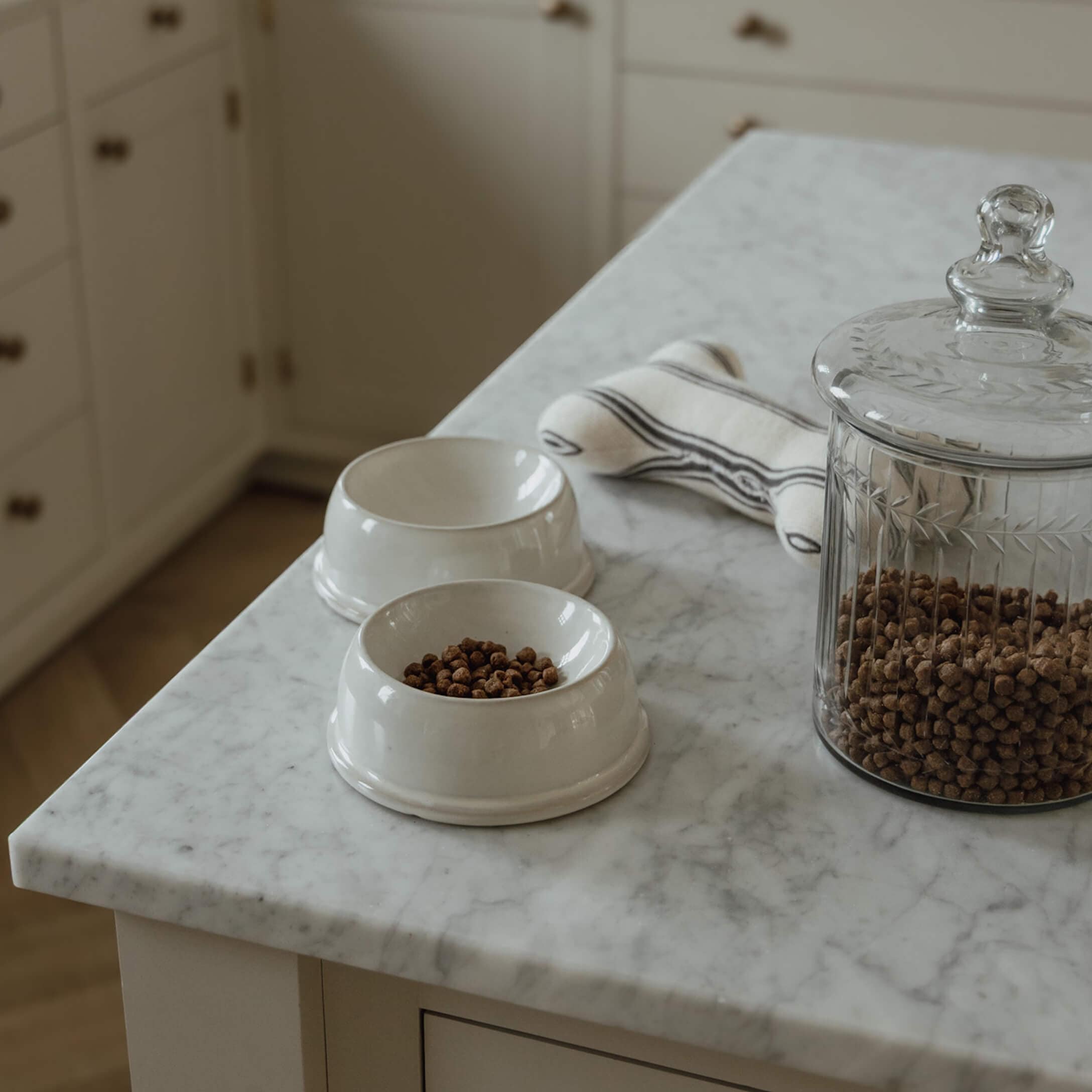 Deux bols en céramique blanche avec croquettes et un bocal en verre sur un plan de travail en marbre