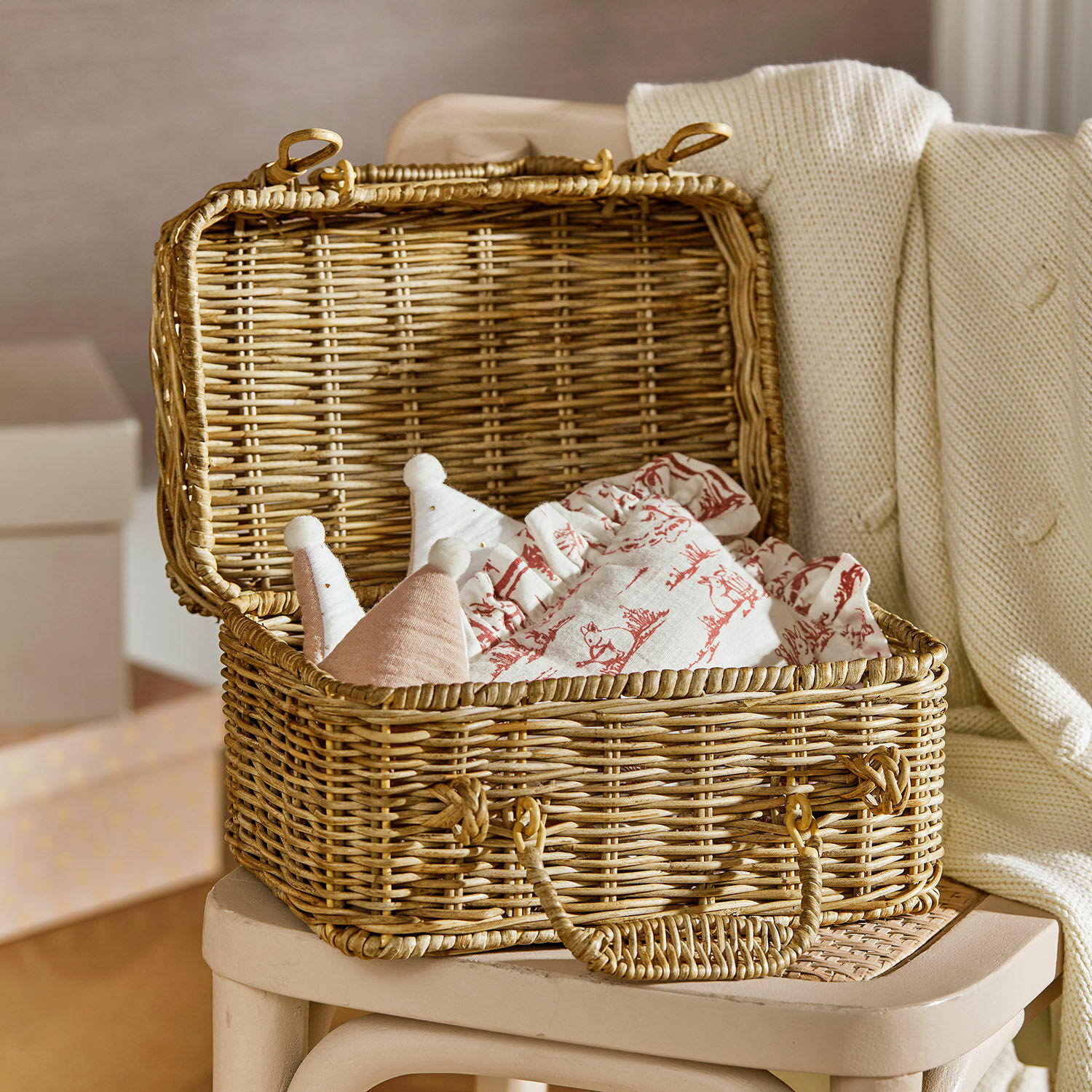 Malle Fableton Panier en osier avec couvercle sur une chaise, rempli de tissus à motifs rouges et de jouets doux