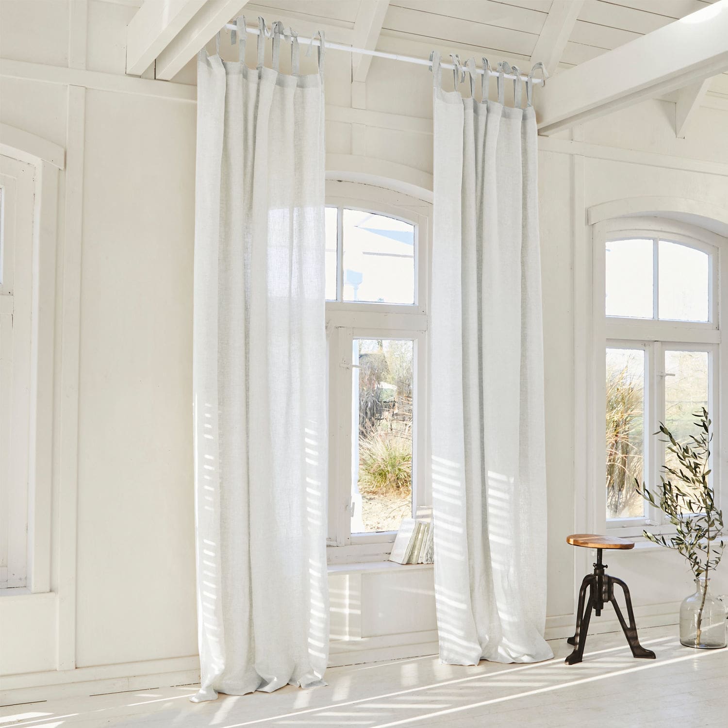 Voilage Monthelie Rideaux en lin blanc suspendus dans une pièce lumineuse avec sol en bois et tabouret en bois