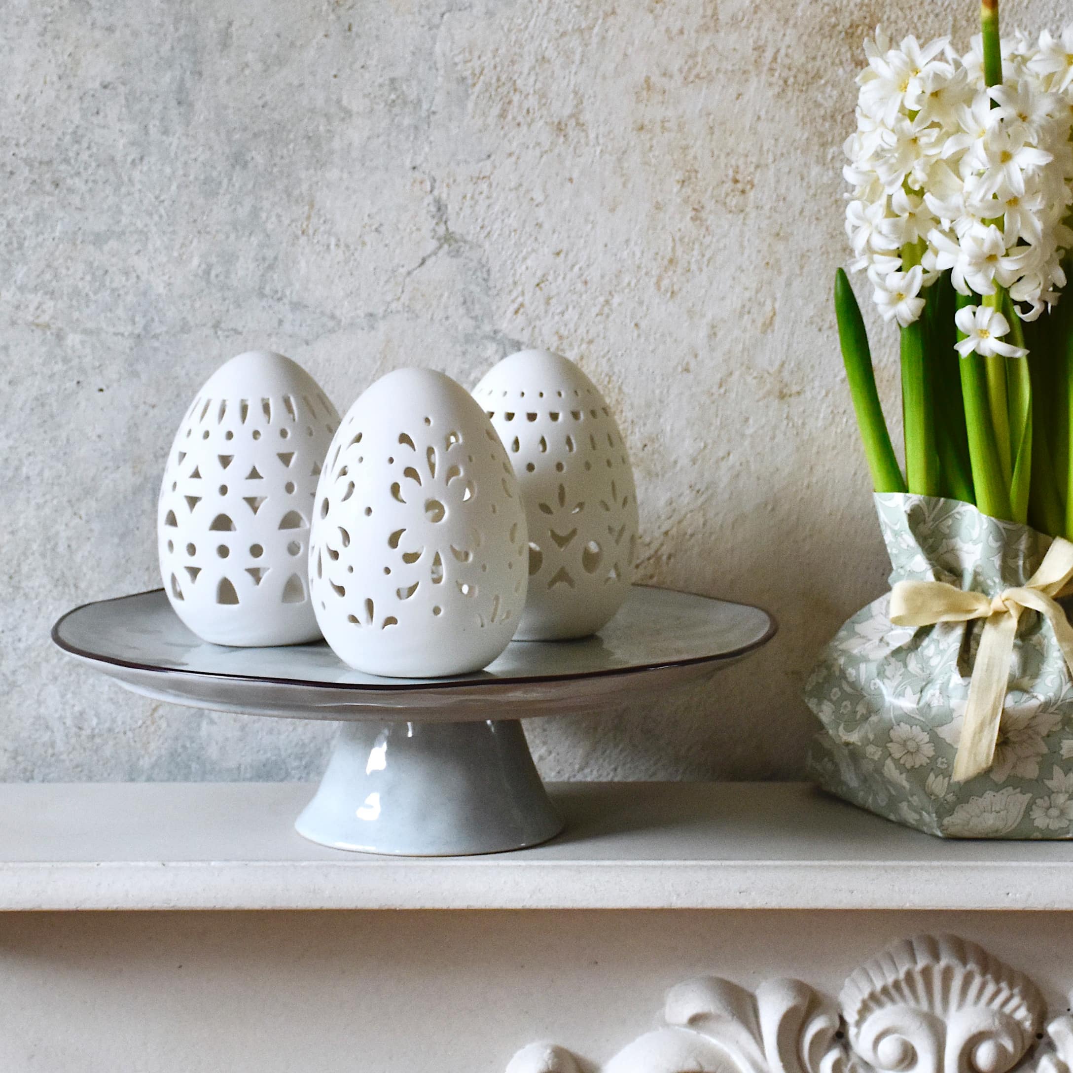 Œufs en céramique blanche ajourée sur un plat gris à côté de fleurs blanches dans un emballage pastel