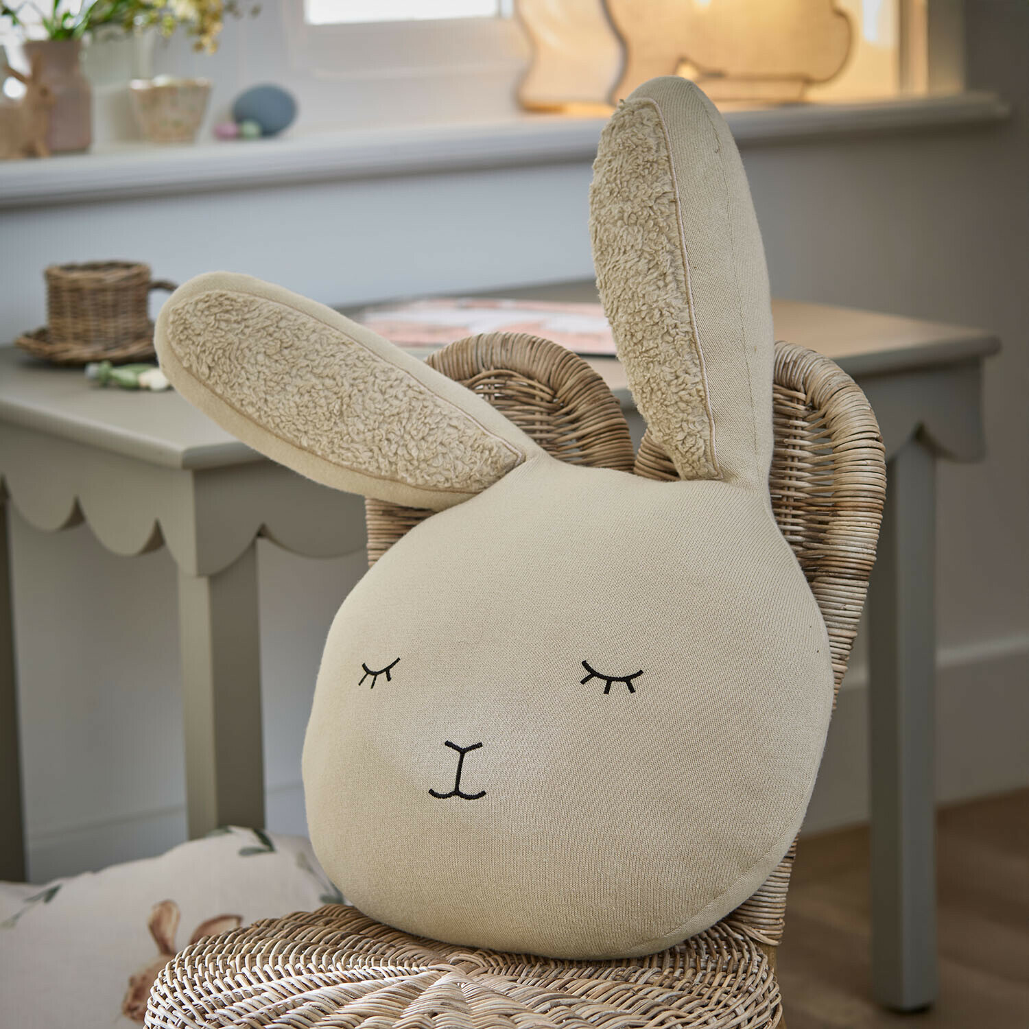 Coussin Fuzzletop Coussin beige en forme de lapin en tissu doux posé sur une chaise en osier dans une chambre d'enfant