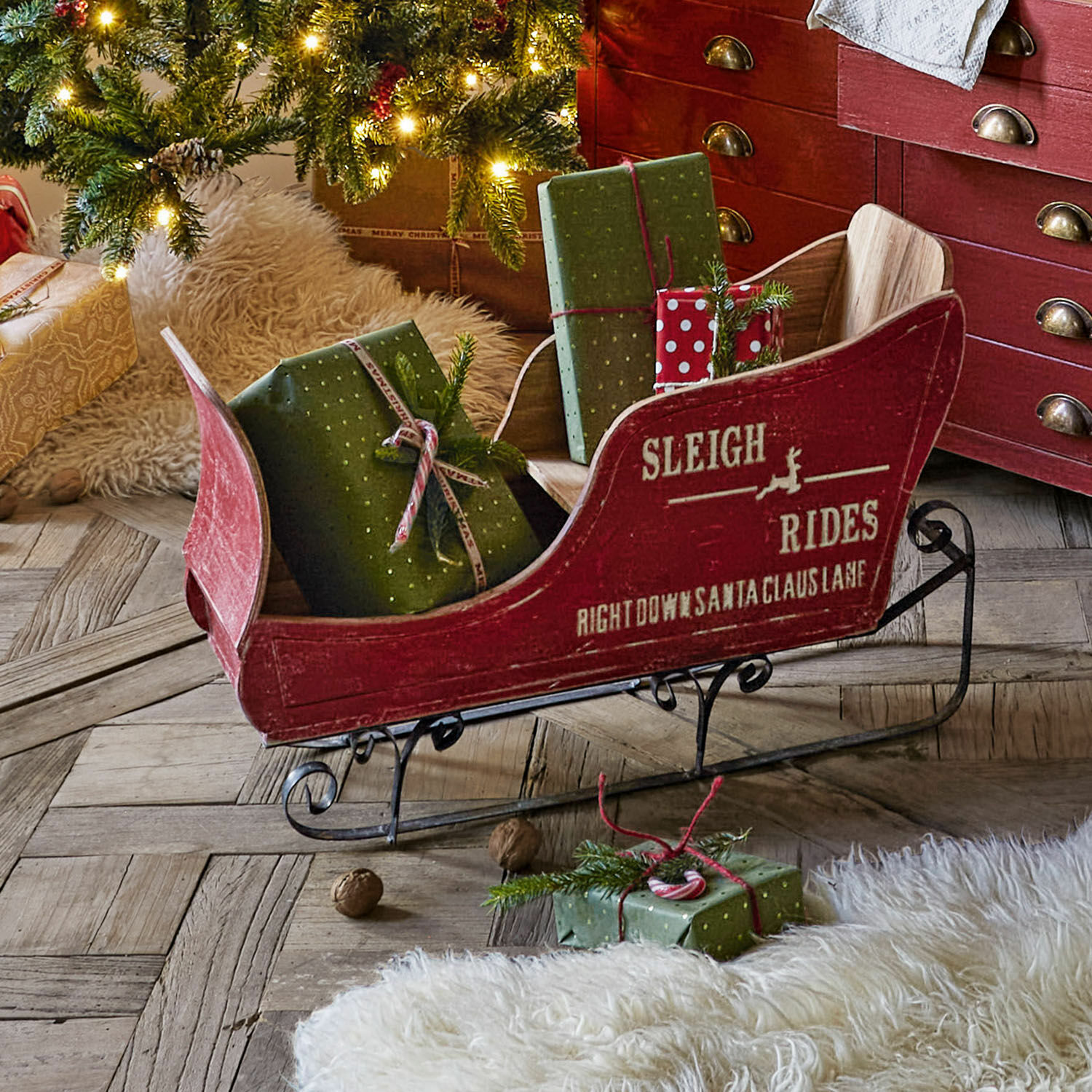 Decoratieve slee Maélys Rode houten slee met metalen runners en groene cadeaus op houten vloer bij versierde kerstboom