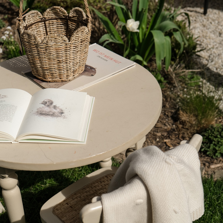 Table ronde beige en extérieur avec panier en osier et livres ouverts dans un jardin ensoleillé