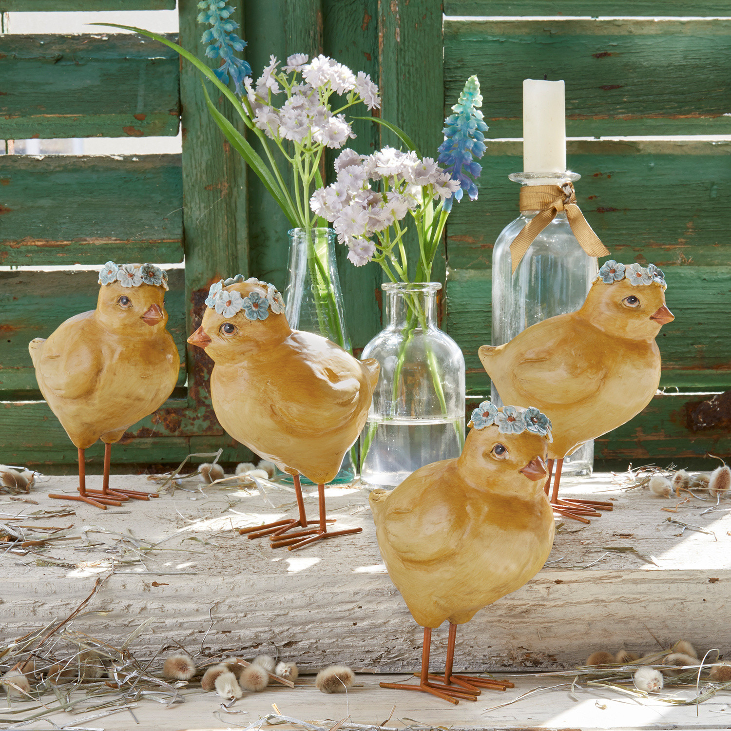 Quatre poussins décoratifs jaunes en matériau avec couronnes de fleurs bleues sur fond de bois vert vieilli