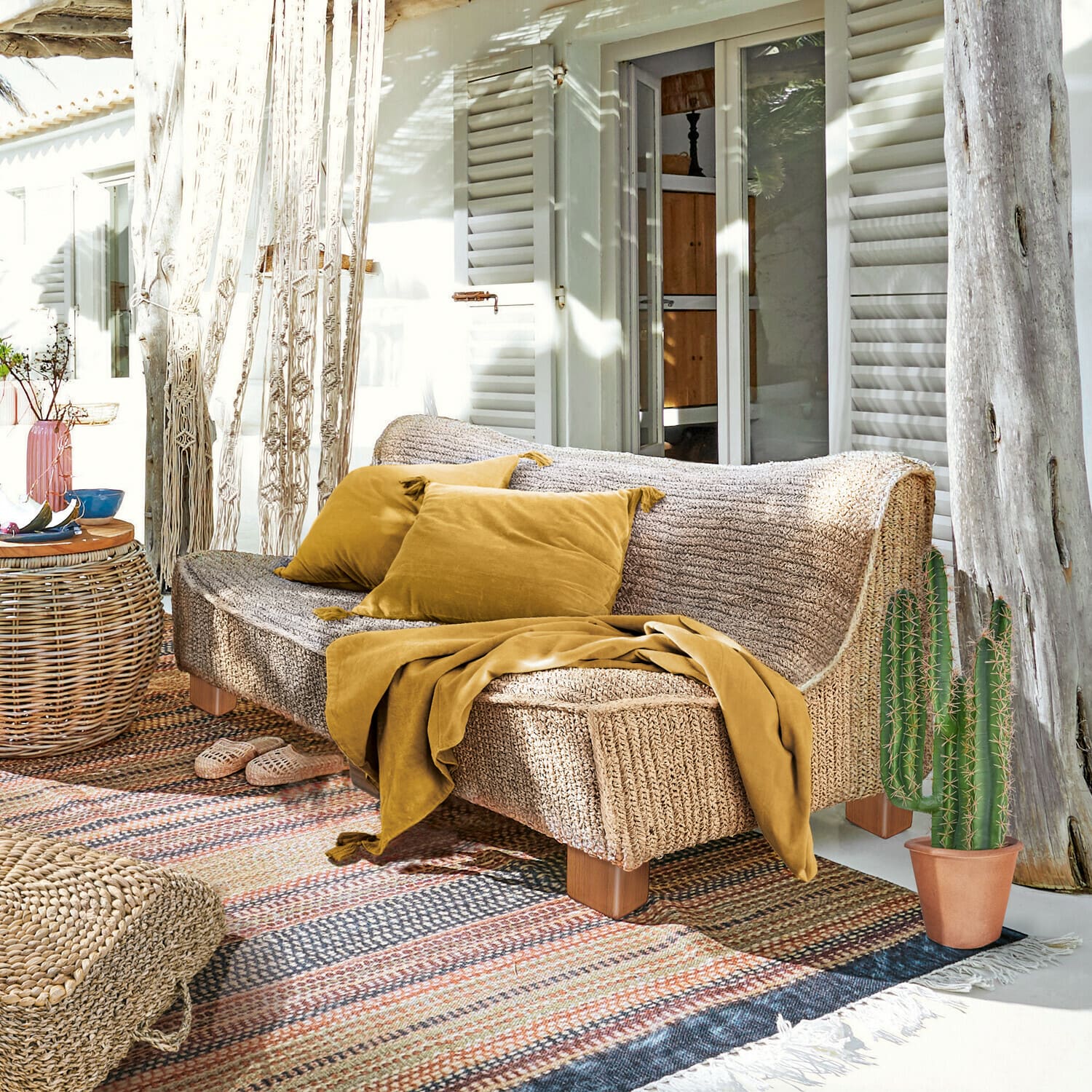 Bank Bocage Bank van gevlochten materiaal in natuurlijke kleur met mosterdkleurige kussens en deken op zonnig terras