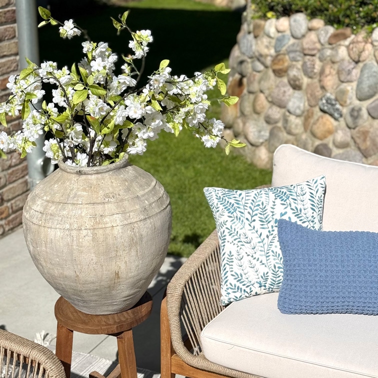 Grote beige keramische vaas met witte bloemen op houten standaard naast bank met kussens in tuin
