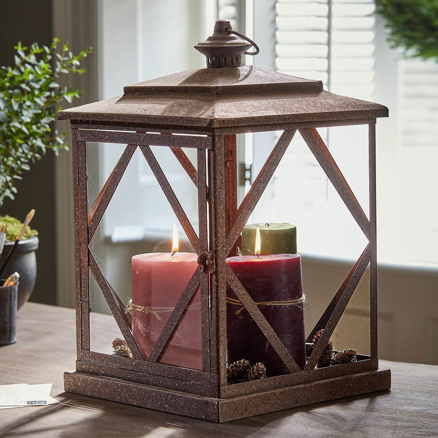 Decoratieve metalen lantaarn in roestkleur met drie kaarsen en dennenappels op tafel