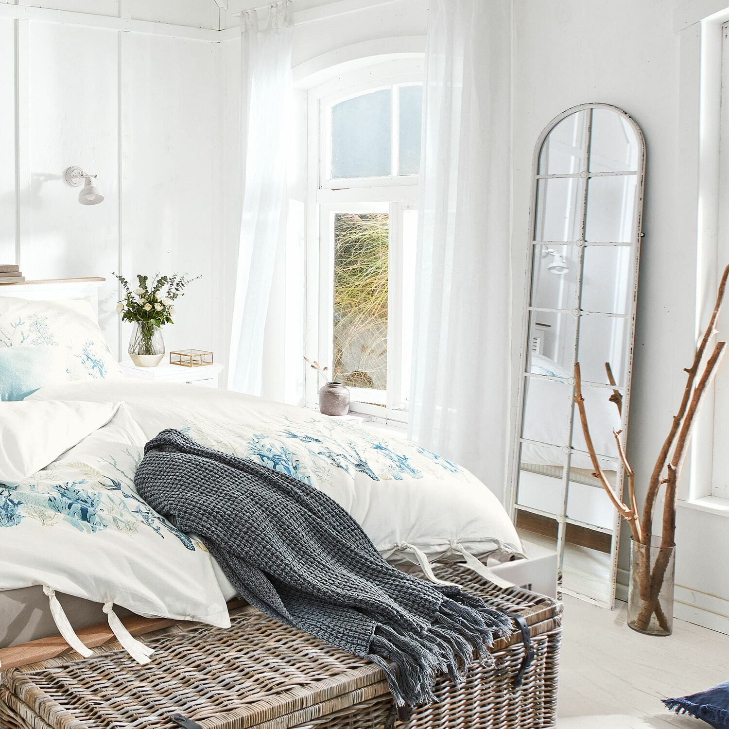 Miroir Westmor Chambre lumineuse avec literie blanche à motif bleu et plaid gris en tricot sur coffre en osier