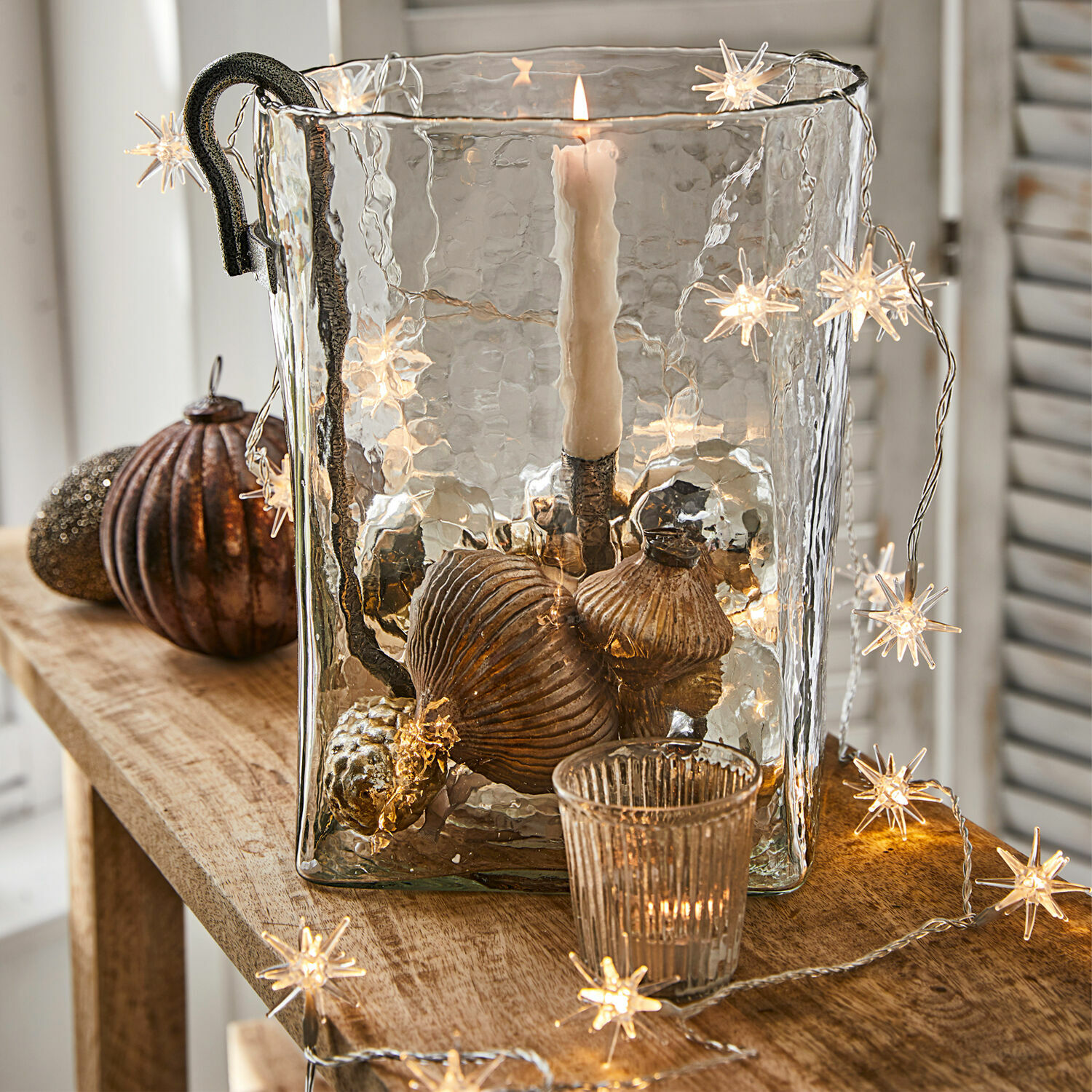 Guirlande lumineuse Nelisse Bougeoir en verre avec poignée en métal et décorations cuivre sur table en bois avec guirlande lumineuse étoile